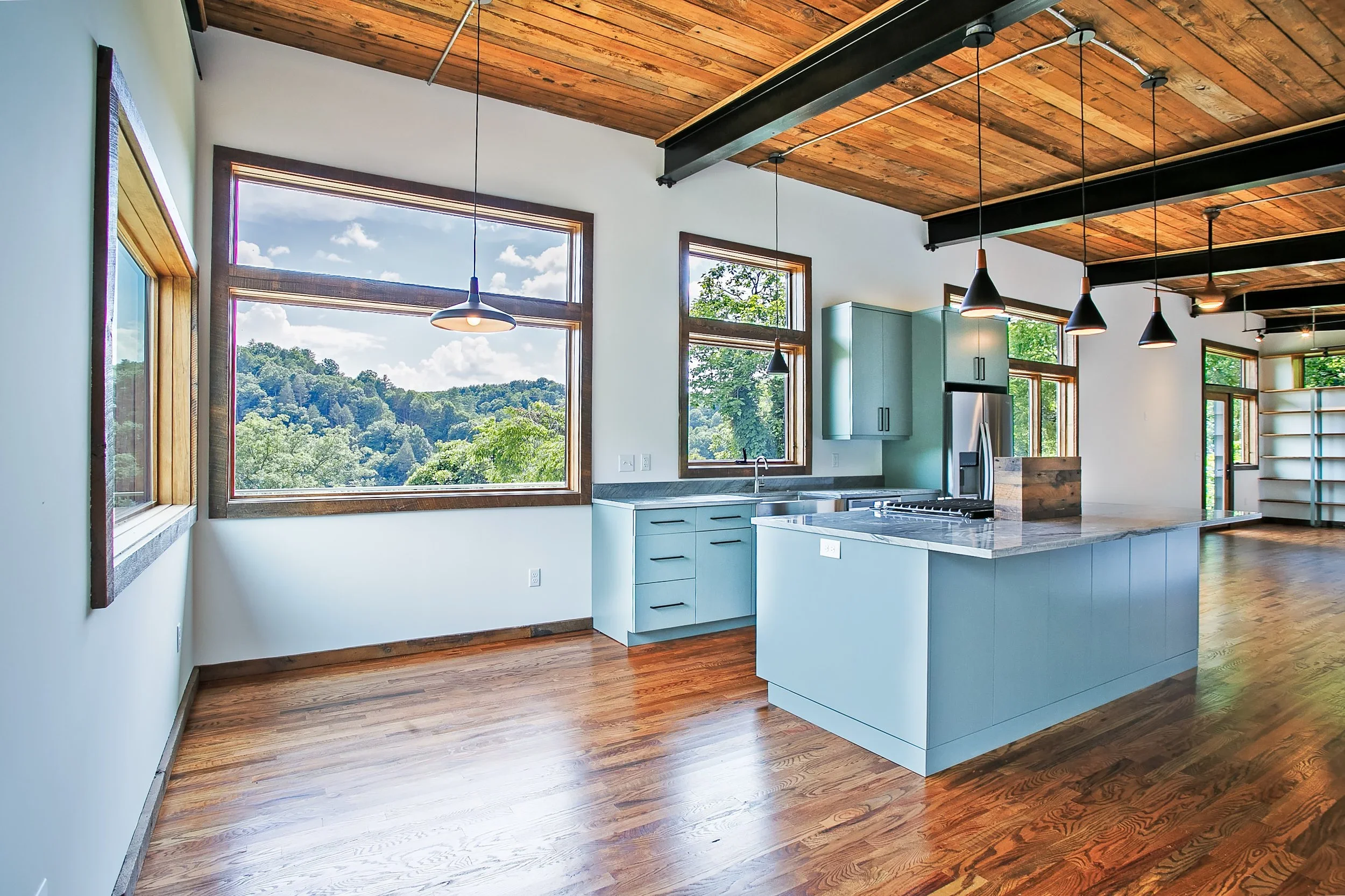 Modern kitchen with large windows, wooden ceiling, hardwood floors, and blue cabinetry.