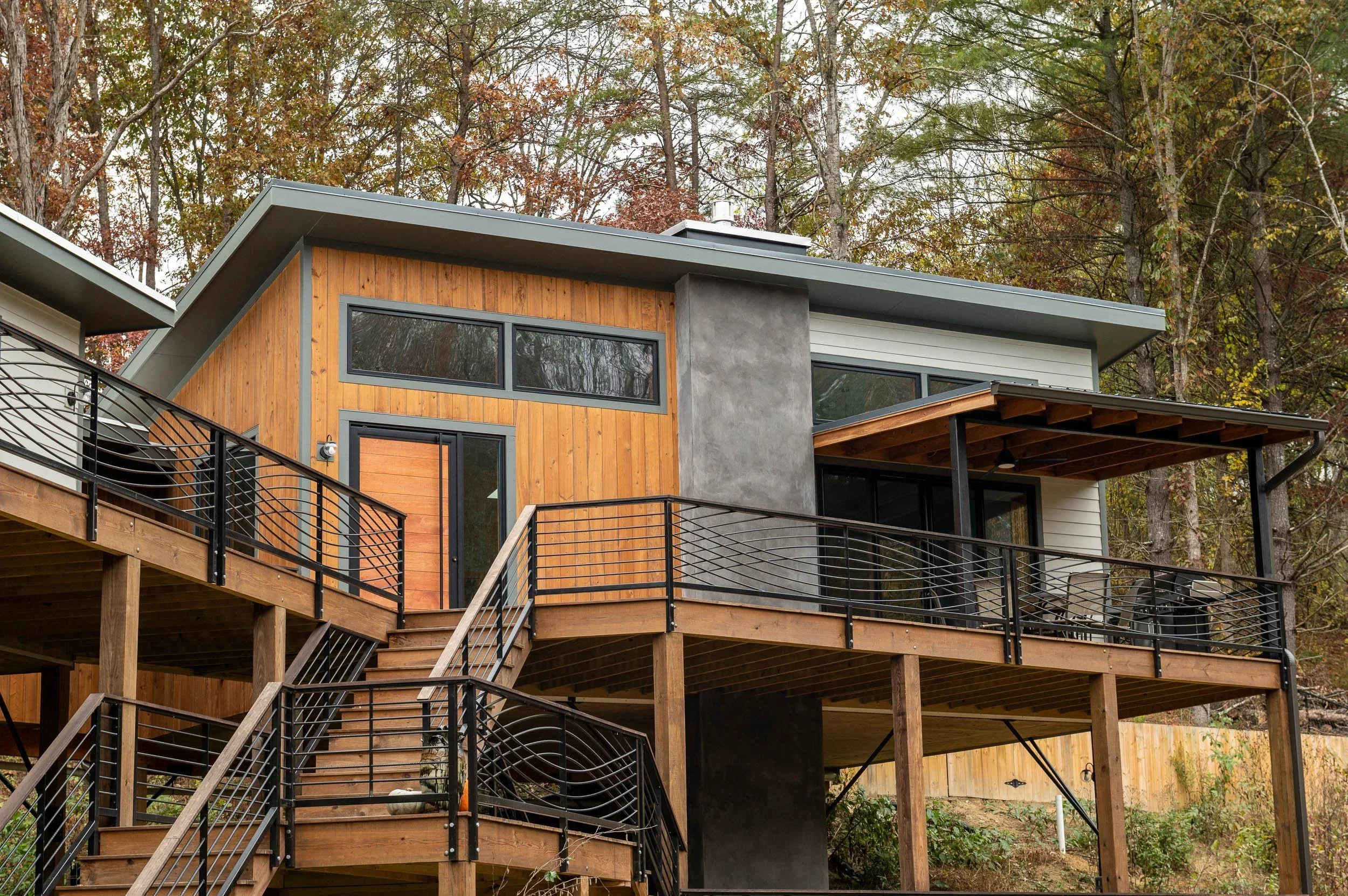 Modern house with wooden and concrete exterior, metal railings, and a large deck overlooking a wooded area.