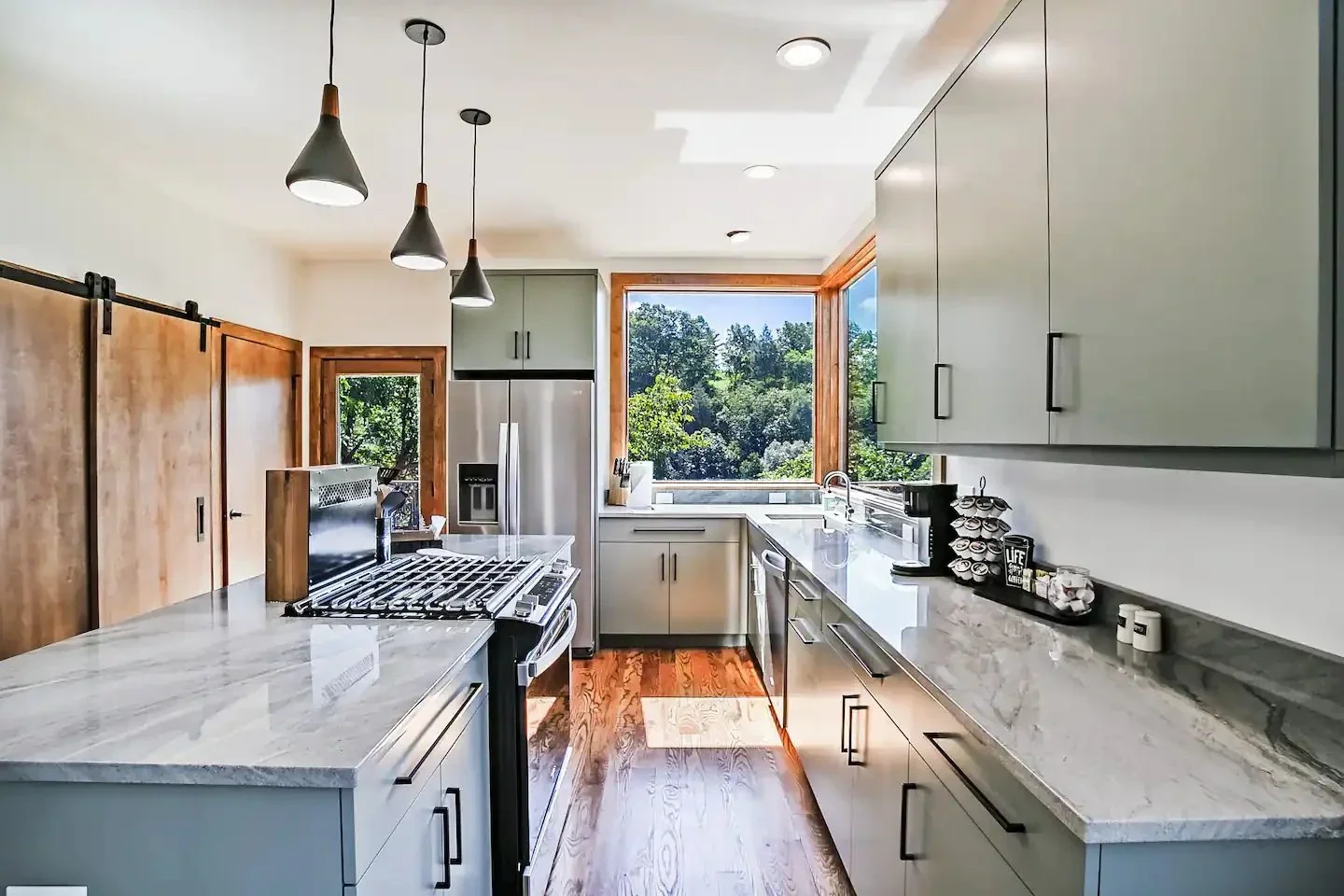 Modern kitchen with white cabinets, marble countertops, large window showing a green outdoor view, and pendant lights hanging from the ceiling.