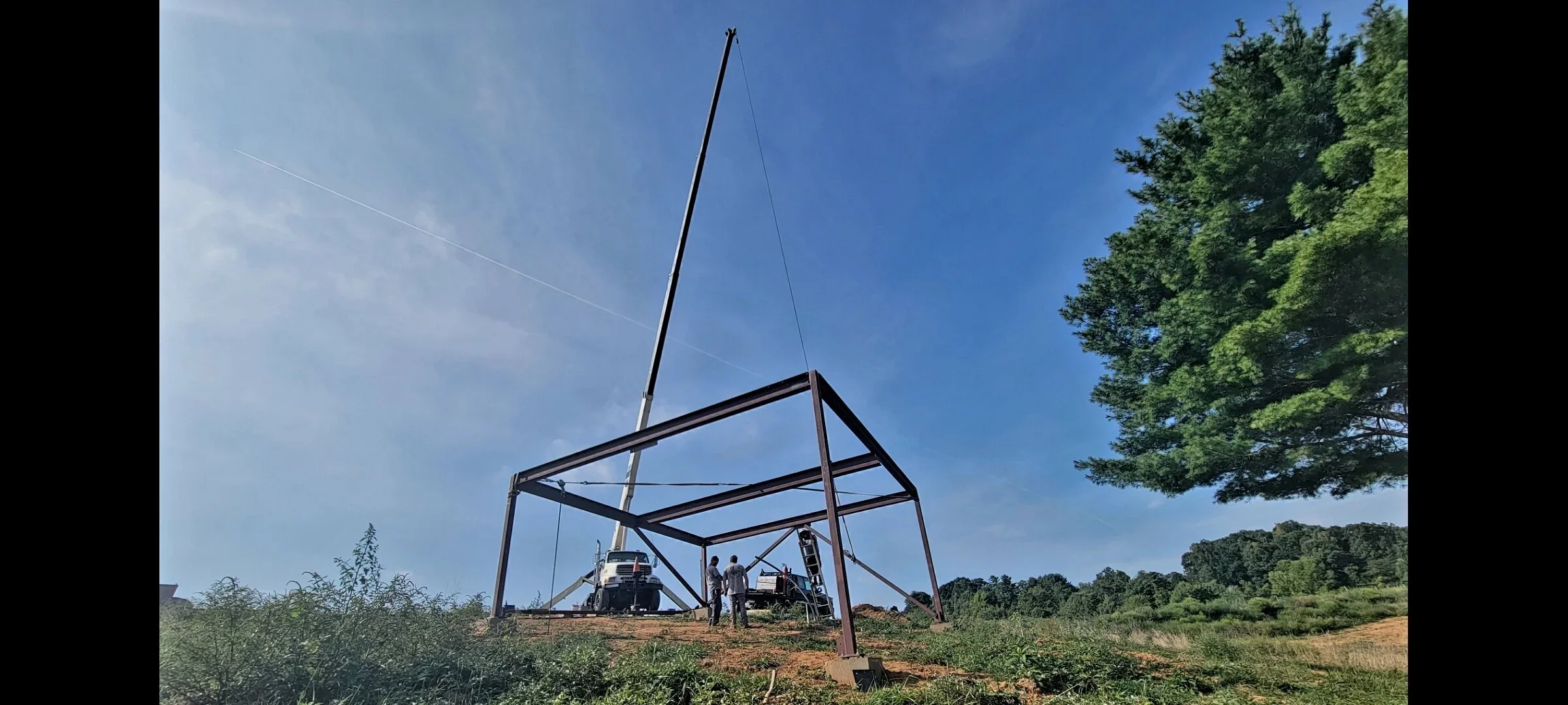 People working on a large antenna tower in an outdoor area with trees and clear blue sky.