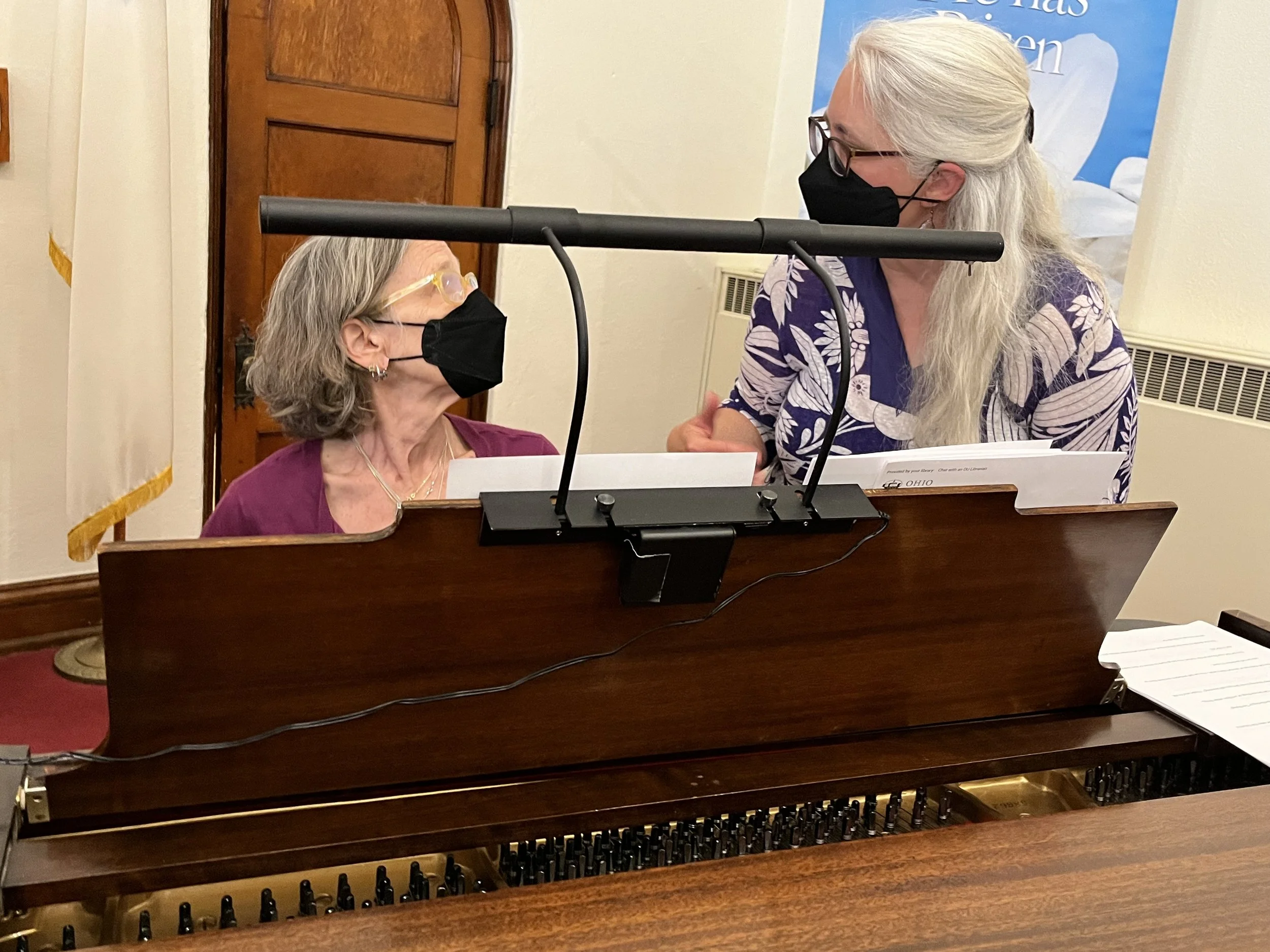 Two women, wearing masks, talk behind a piano.