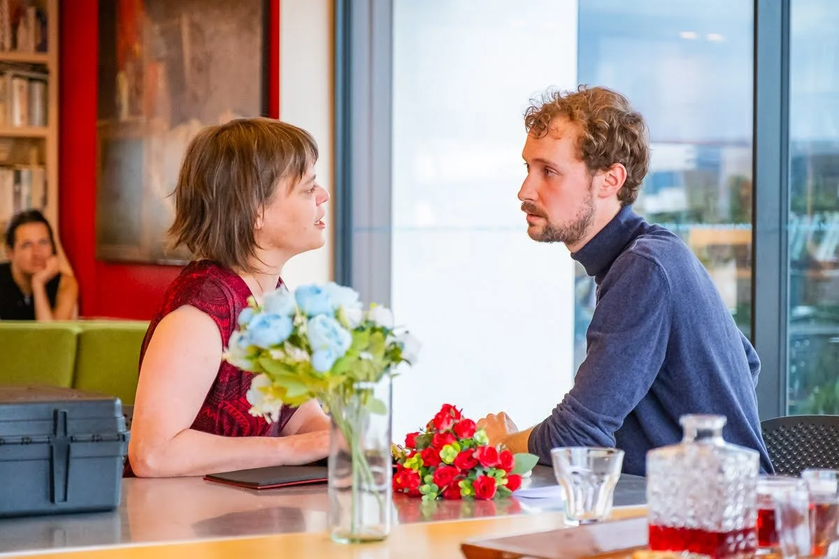 A man and woman sitting across from each other at a table with flowers, engaged in a serious conversation. In the background, a woman is sitting on a green couch, watching them.
