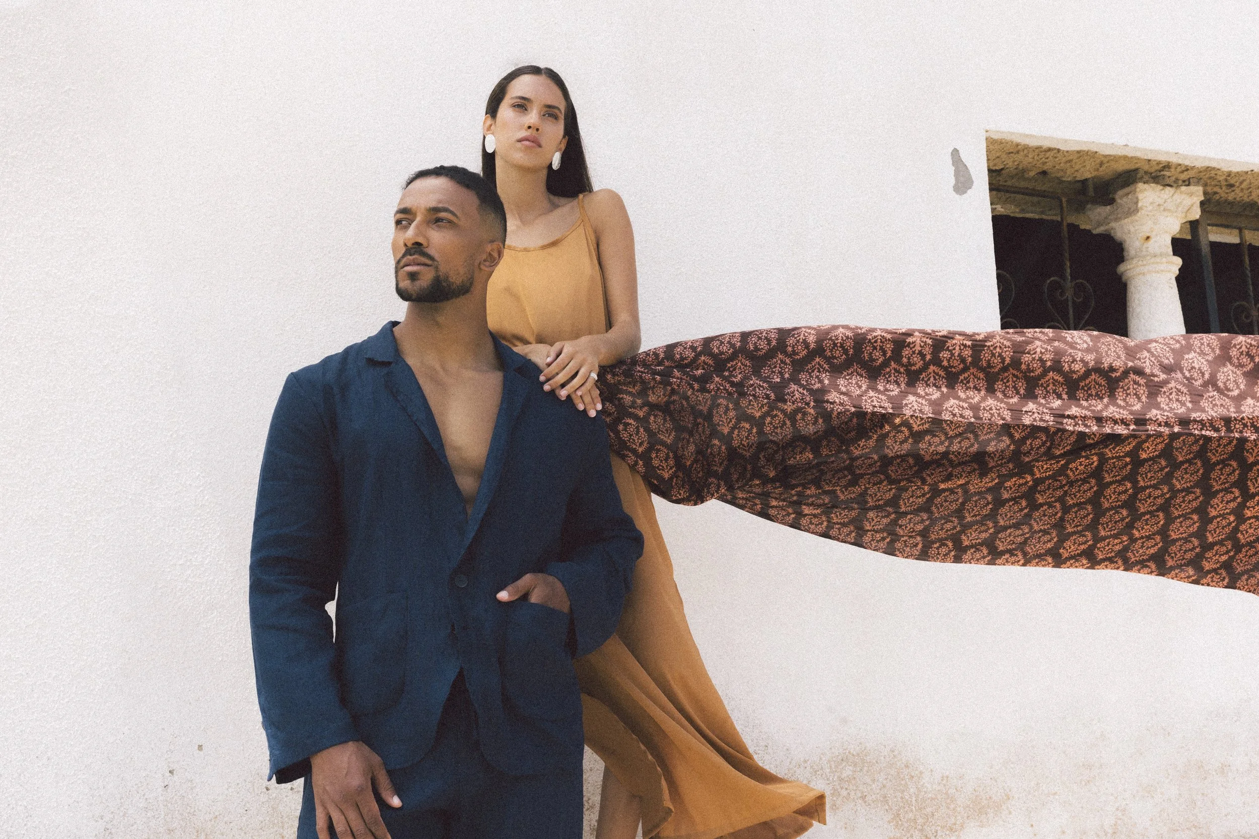 A man and woman standing against a white wall, with the woman leaning on a ledge with patterned fabric.