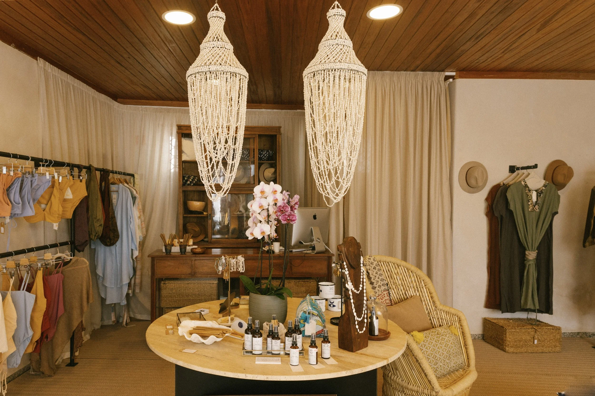 A boutique store interior with clothing racks on the left displaying various colorful garments, a wooden table in the center with skincare products, a potted orchid, and jewelry, a wicker chair with cushions on the right, a wooden sideboard and display cabinet in the background, hanging hats and a dress on the wall, and two large woven pendant lights hanging from the wooden ceiling.