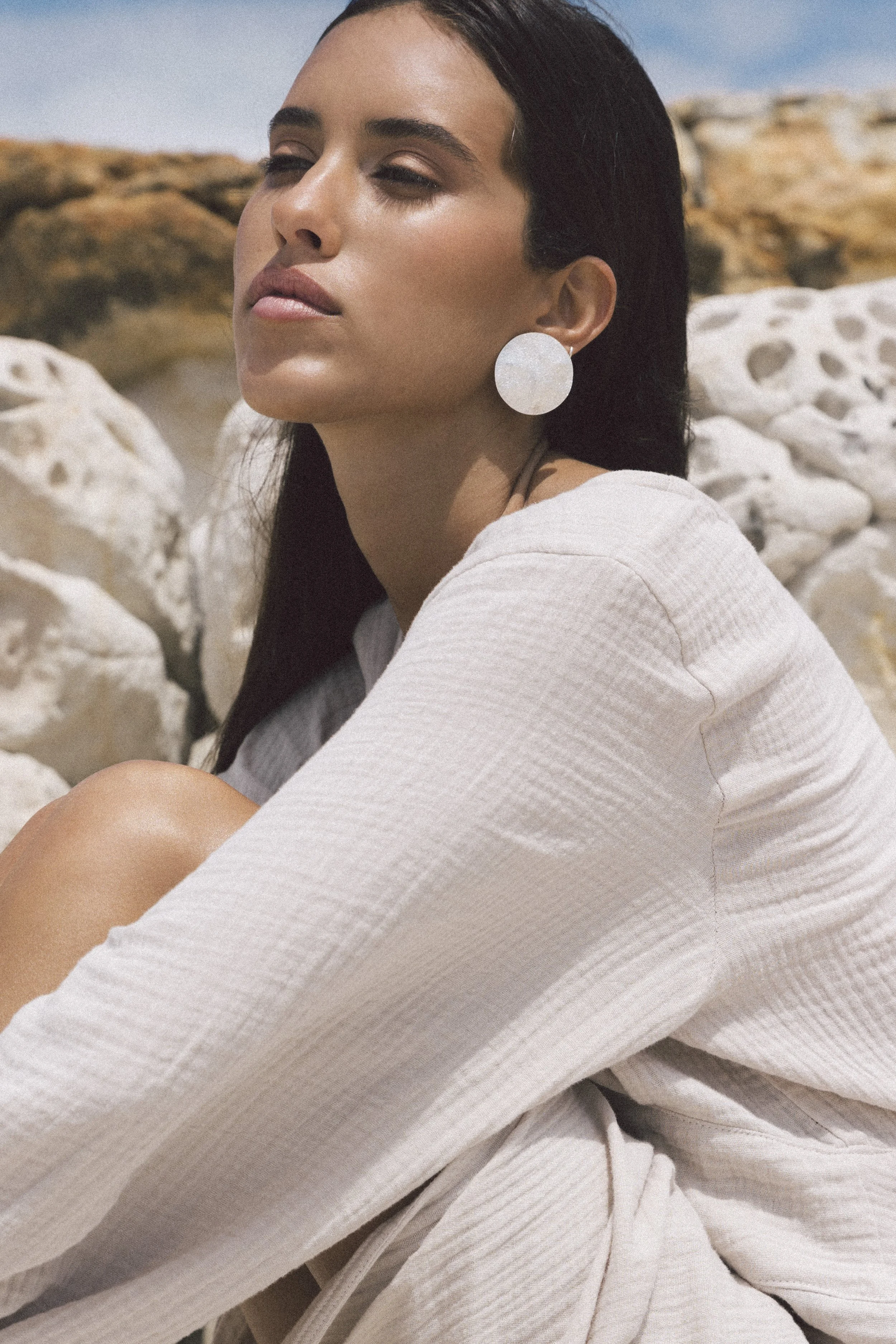 A woman with dark hair wearing large round earrings sits against a rocky background with white stones, outdoors on a sunny day.