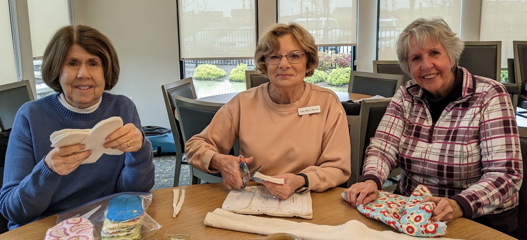 Three women sitting at a table engaging in a craft activity with fabric pieces and sewing supplies.