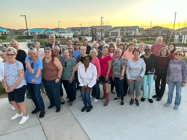 Group of diverse adults posing outdoors, standing closely together, smiling with a background of houses and a sunset.