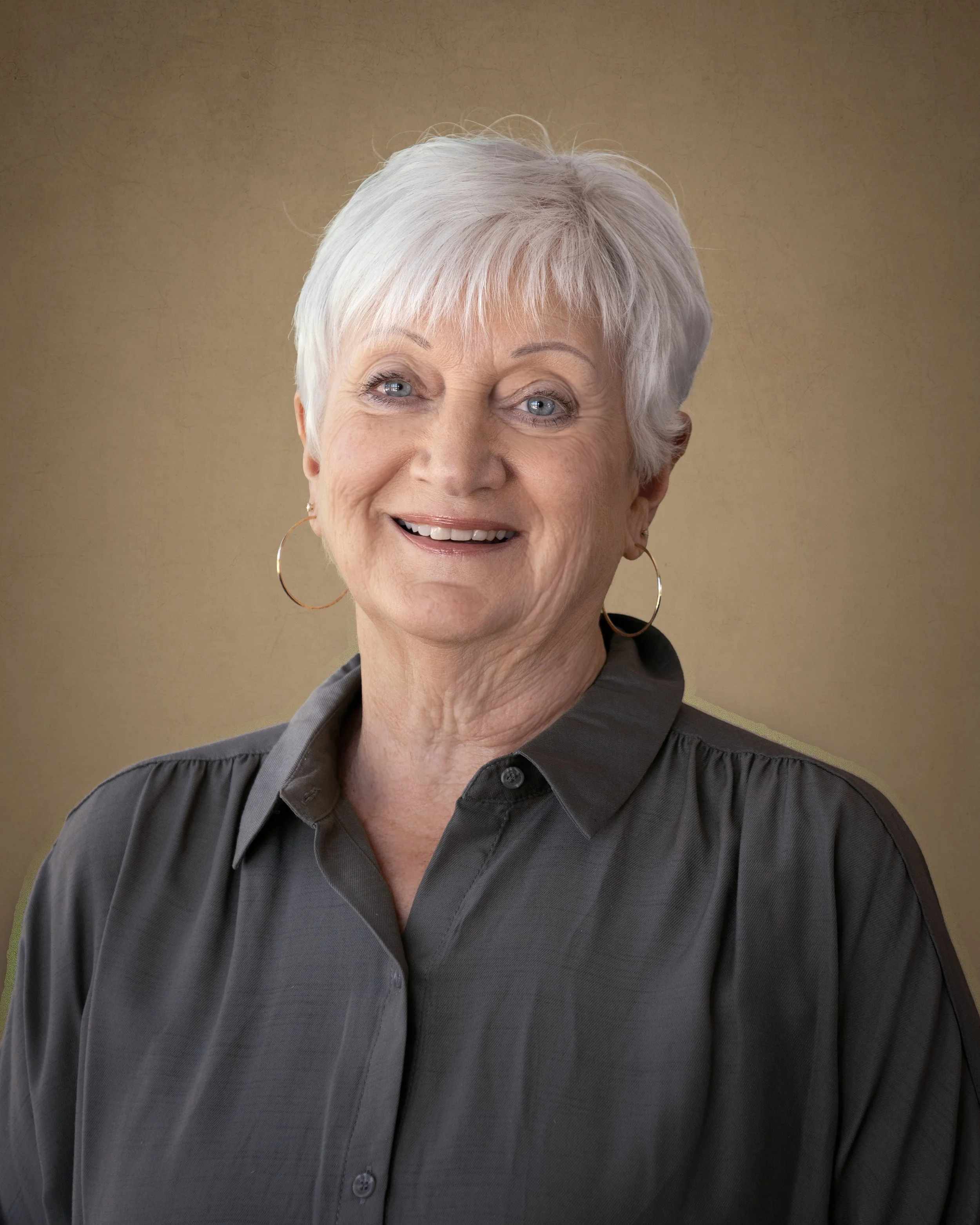 Elderly woman with short white hair and hoop earrings smiling, wearing a dark gray button-up shirt, against a plain background.