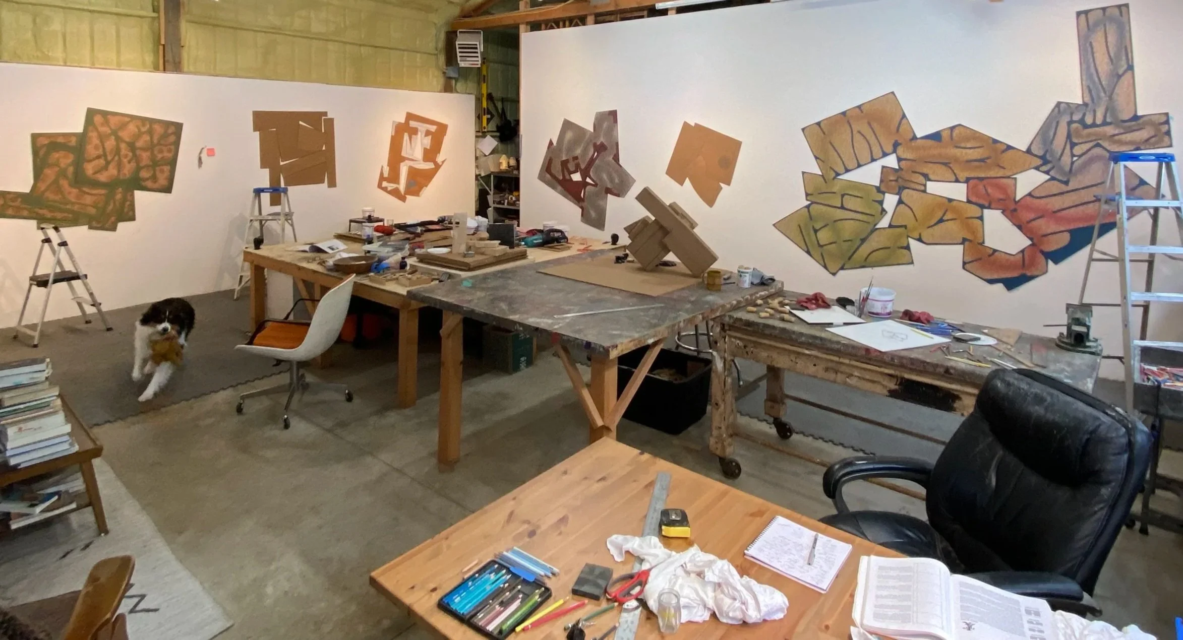 A workshop with various art supplies and three abstract mural paintings in progress on the wall. There is a dog holding a plush toy, several tables with tools, books, and notes, and a stepladder on each side of the wall.