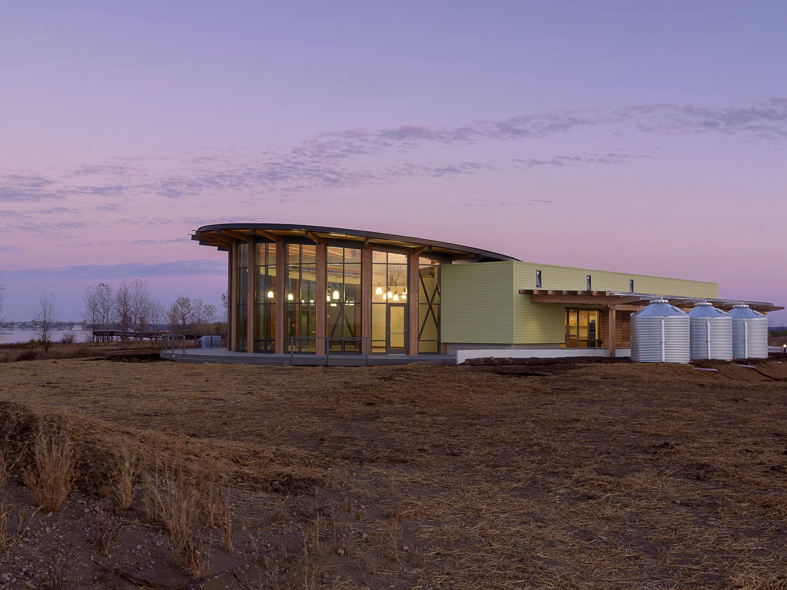 Modern building with large glass windows and metal water tanks on the side, illuminated inside during dusk, set on a barren landscape.