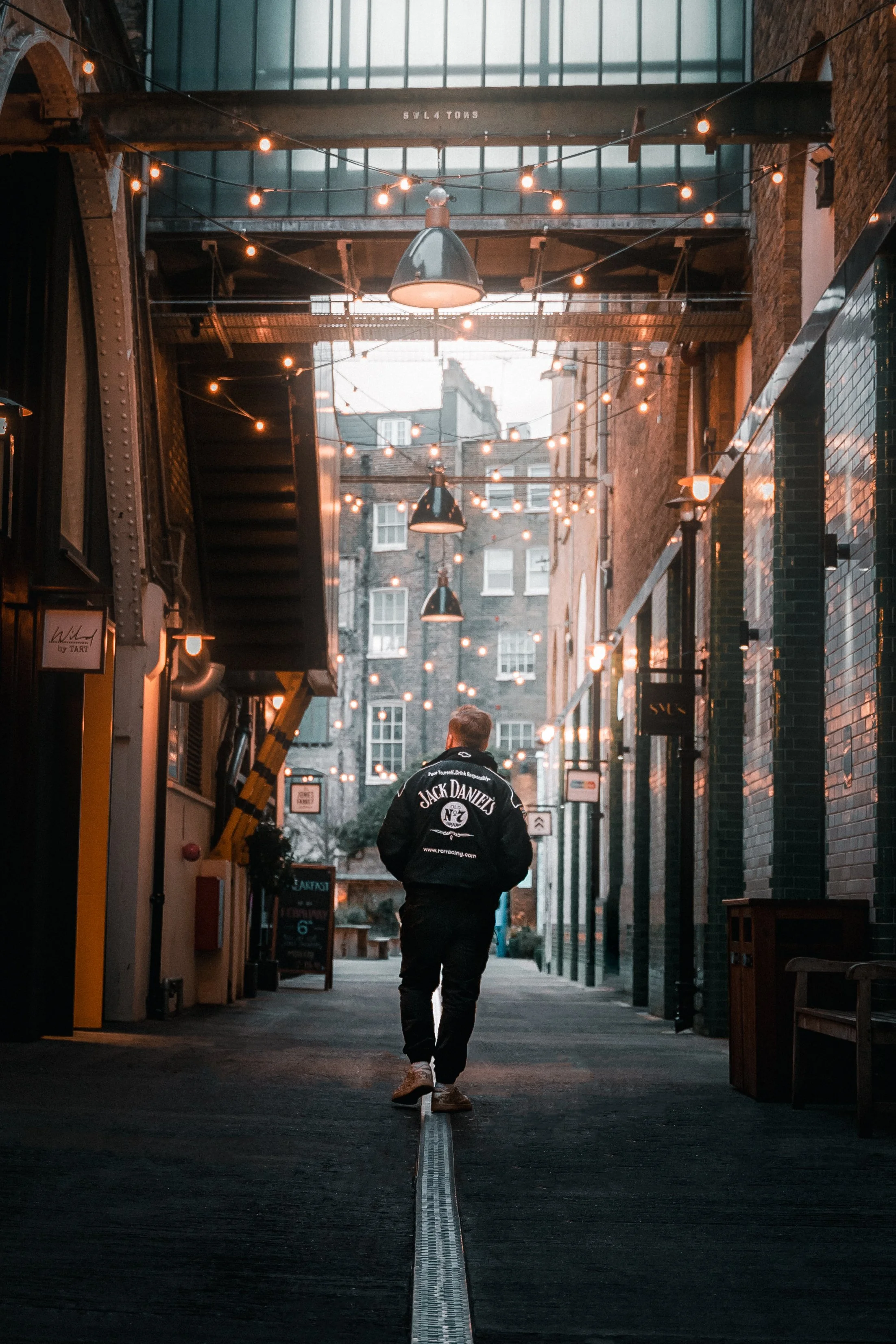 Person walking in an urban alley with string lights overhead, wearing a black Jack Daniels jacket.