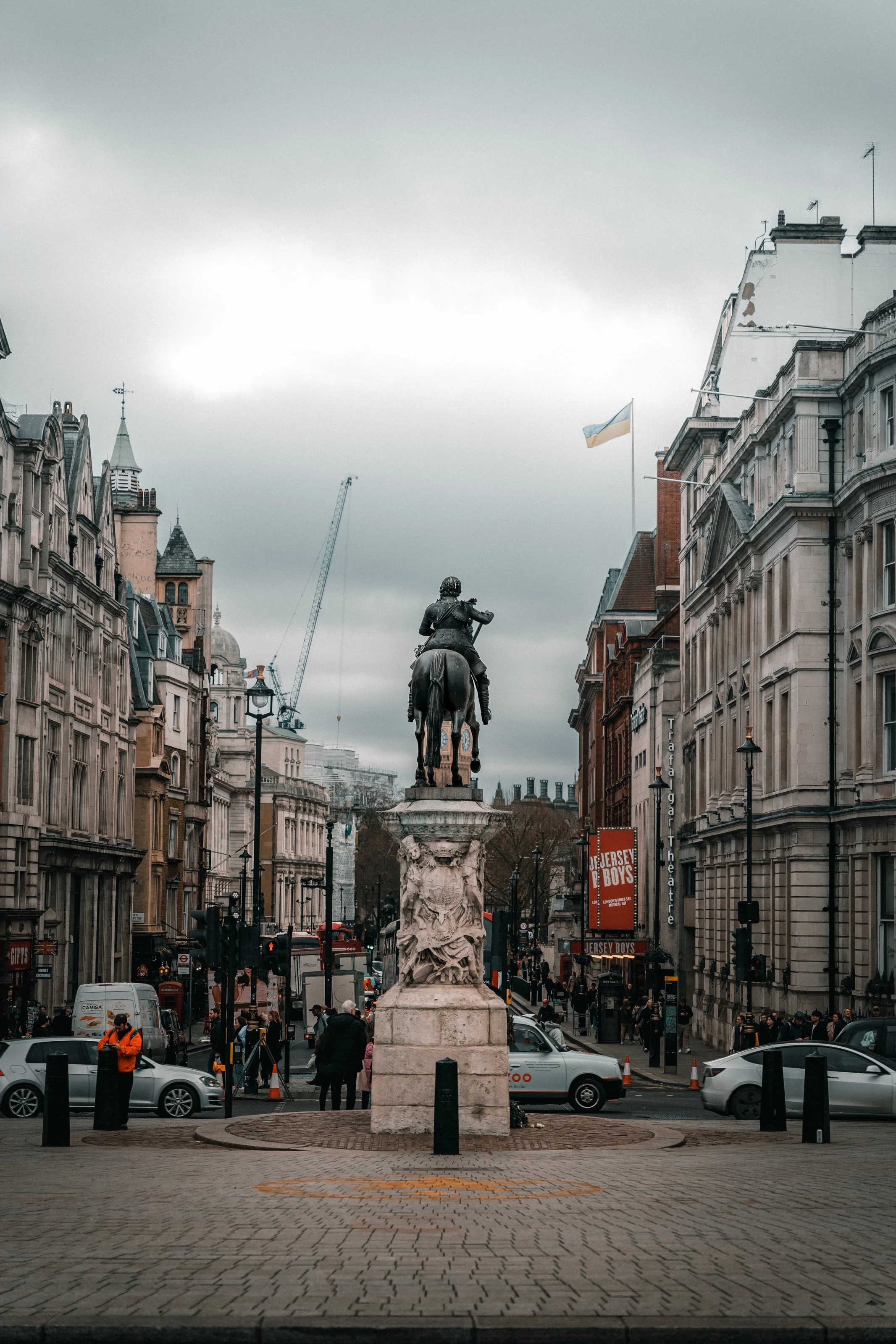 Equestrian statue on a pedestal in a busy urban setting, surrounded by historic buildings and traffic, with a crane and overcast sky in the background.