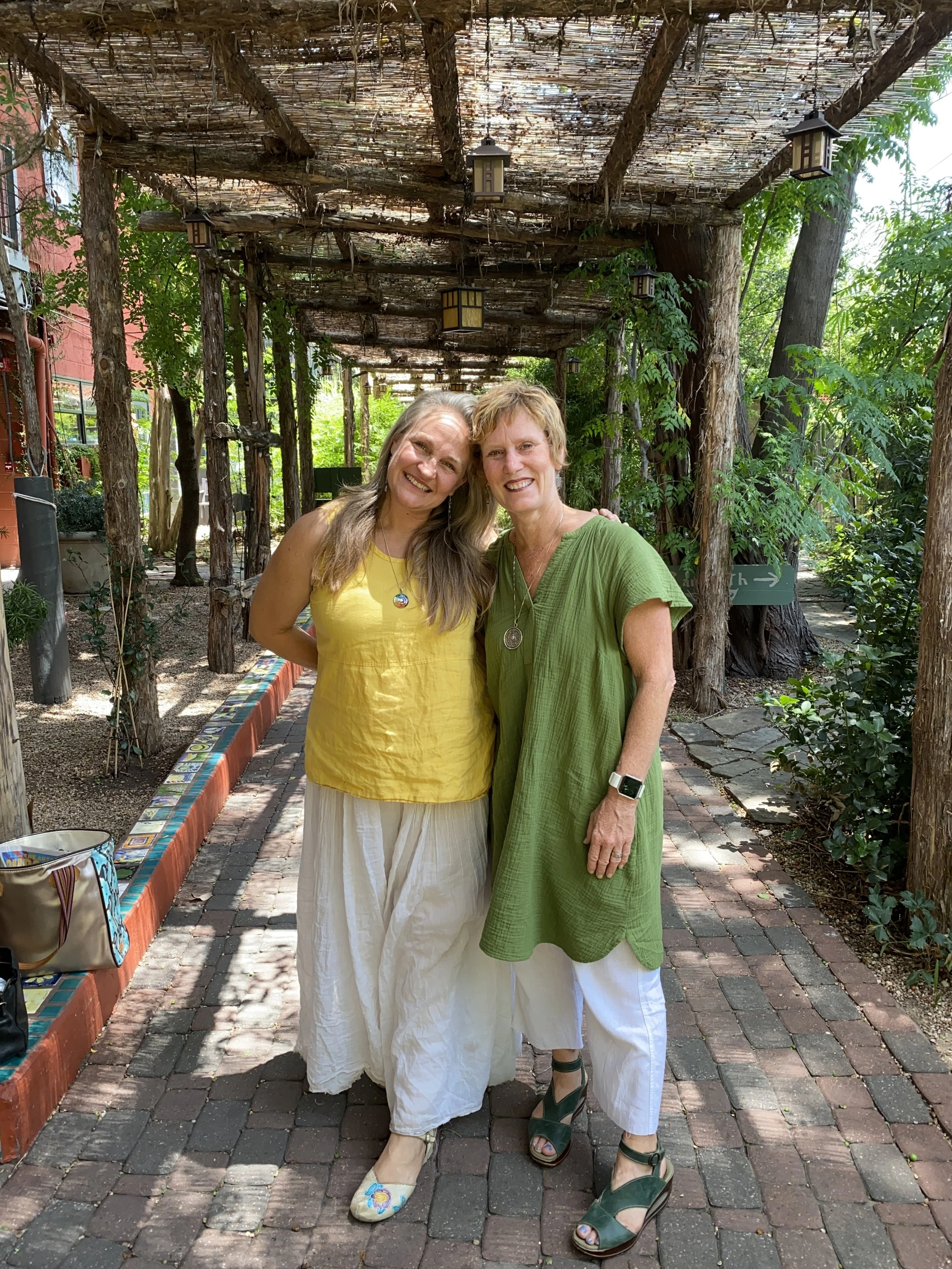Two women smiling and posing closely together in an outdoor garden setting with leafy greenery and a wooden trellis overhead. One woman is wearing a yellow top and white pants, while the other is wearing a green dress and white pants.