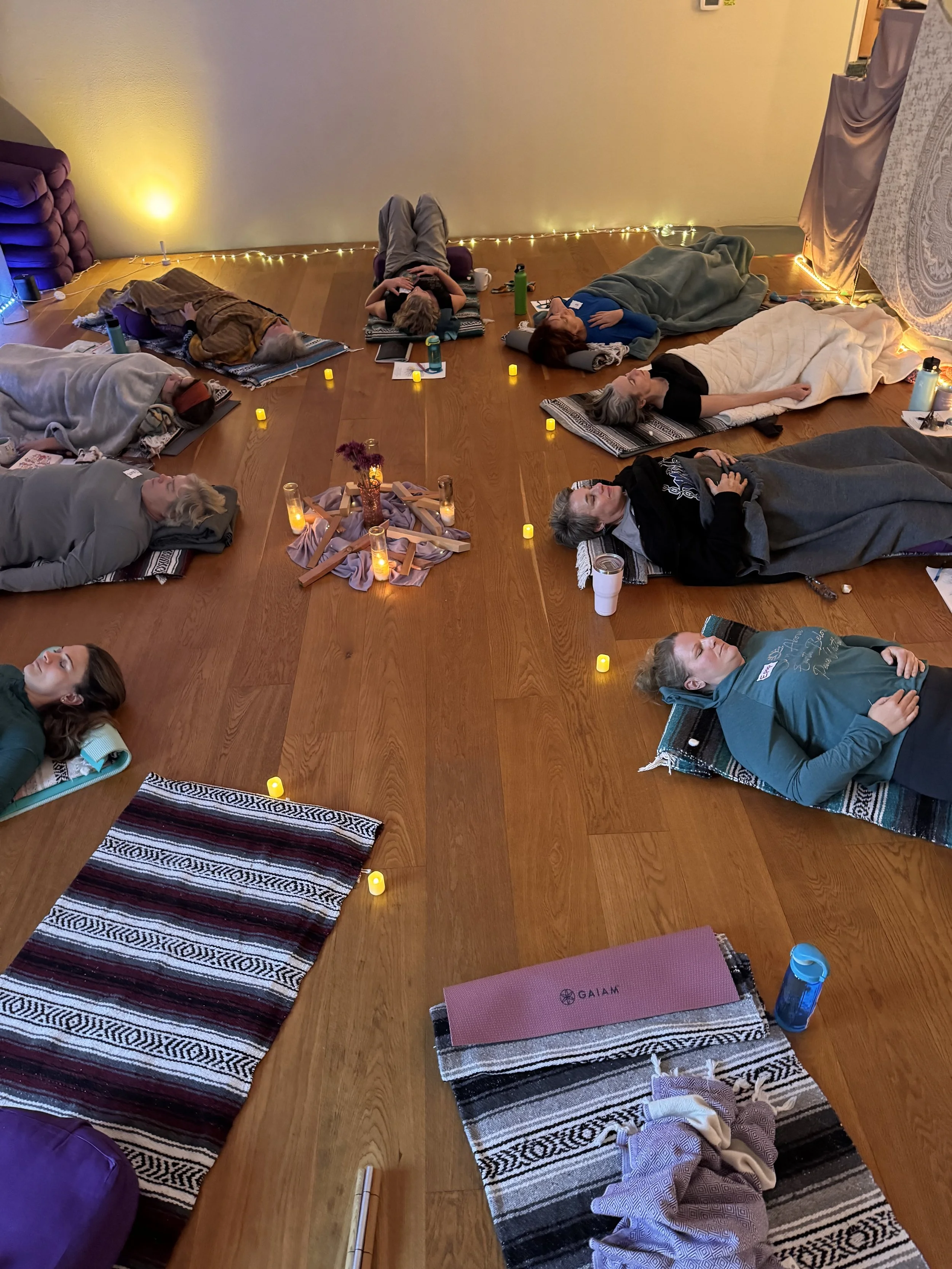 People lying on yoga mats and blankets on a wooden floor, participating in a relaxing meditation session. Small yellow candles are placed around the mats and a small altar with flowers and candles is at the center of the circle. Dim, cozy lighting and fairy lights add to the peaceful atmosphere.