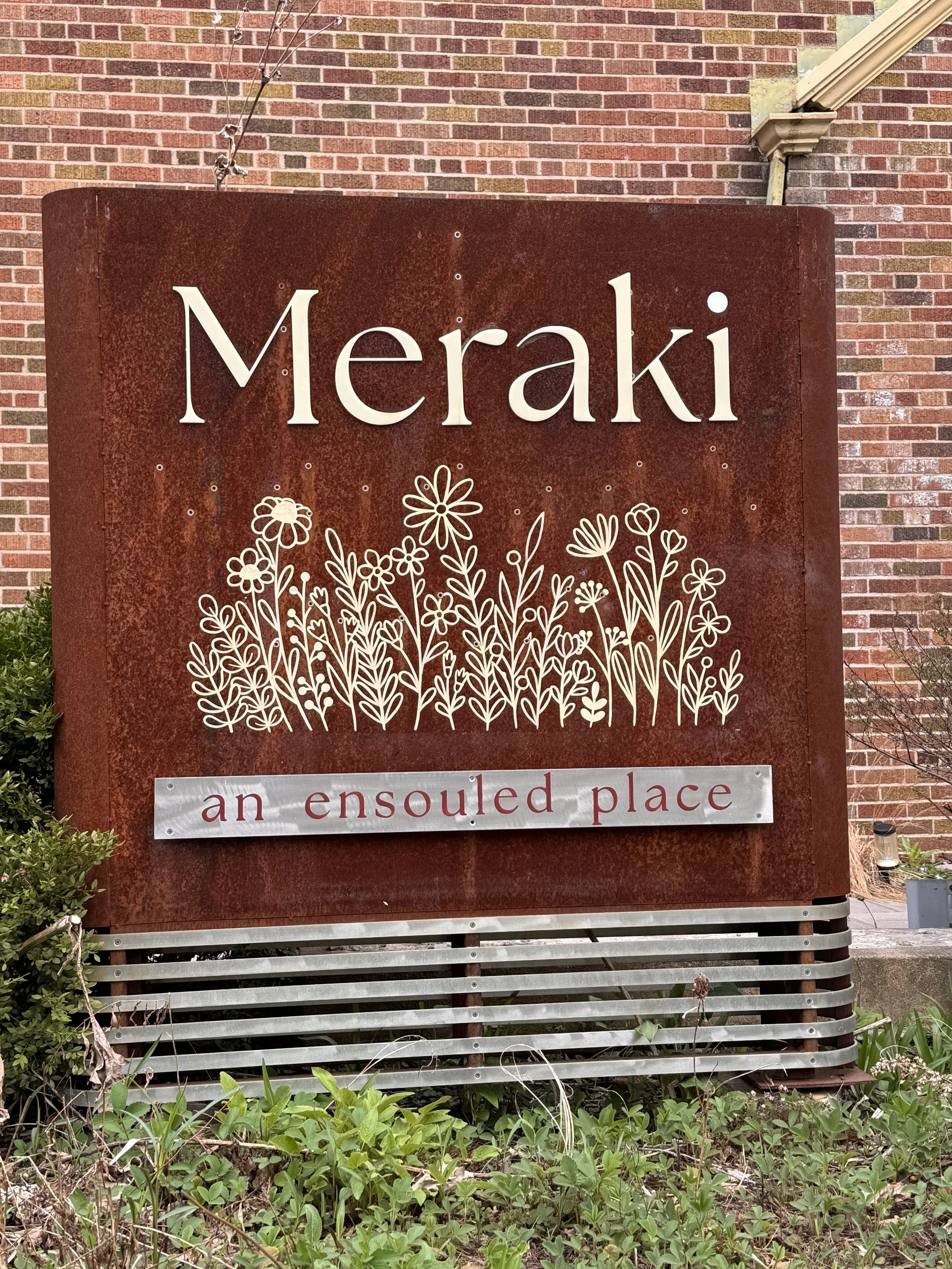 A large reddish-brown metal sign with the word 'Meraki' and an illustration of flowers below it. The sign also has a smaller white text that reads 'an ensouled place'. The sign is in front of a brick wall and some greenery and plants at the bottom.
