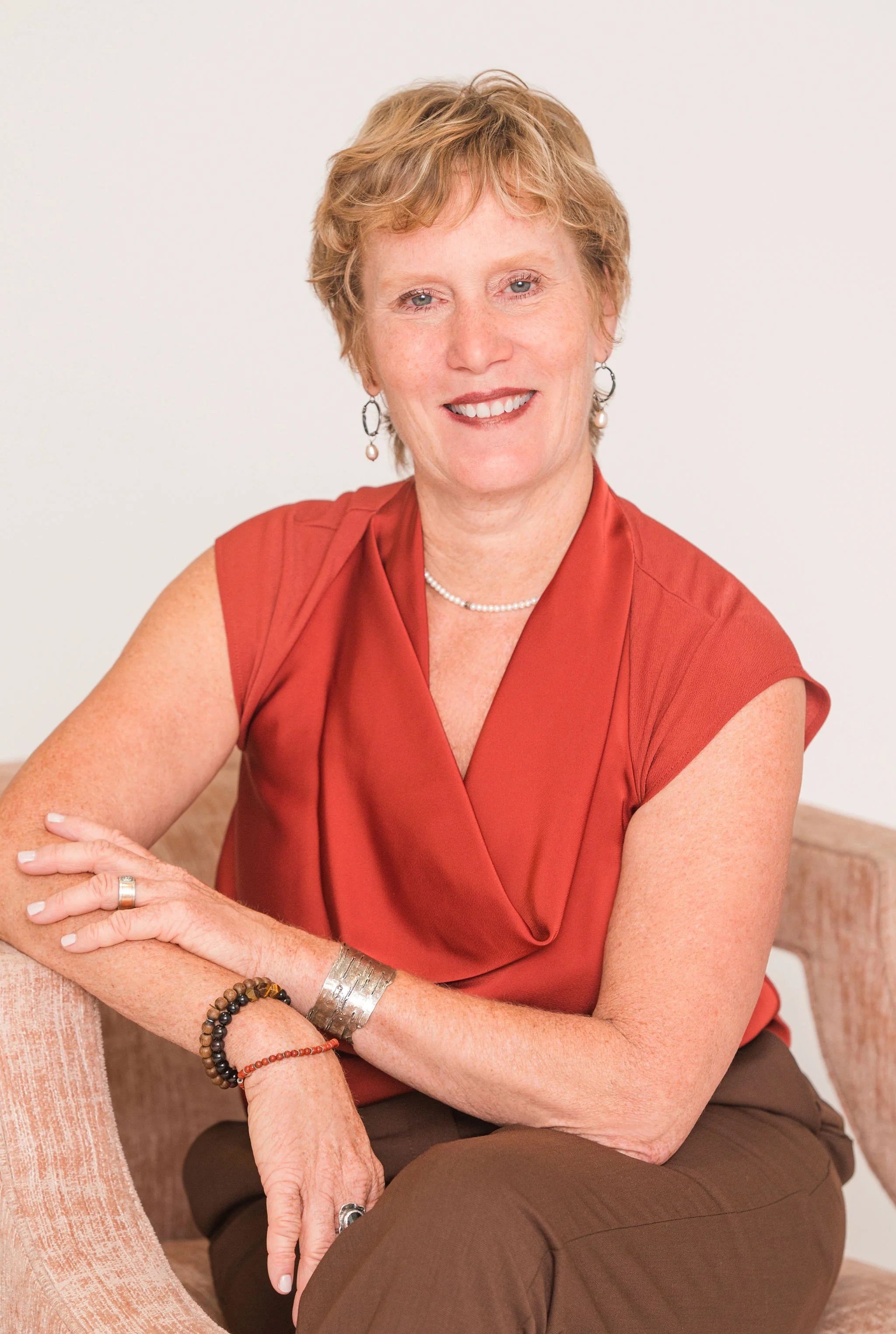 A smiling woman with short, curly blonde hair wearing yellow earrings, a matching yellow blouse, a necklace, and a bracelet while sitting on a black couch. She has blue eyes and is seated in a brightly lit room.