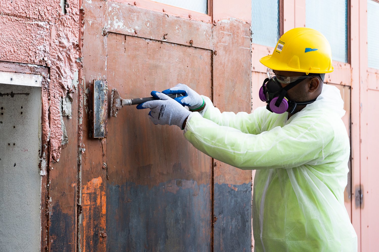 Wearing appropriate Personal Protective Equipment (PPE), a worker scrapes lead contaminated paint. 