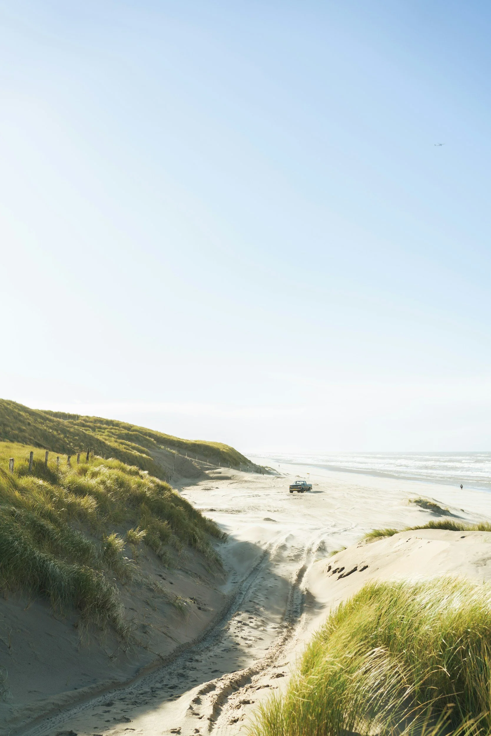 A sandy beach with vehicle tracks, green dunes, and grassy vegetation, under a clear blue sky with distant ocean and a small airplane in the sky.