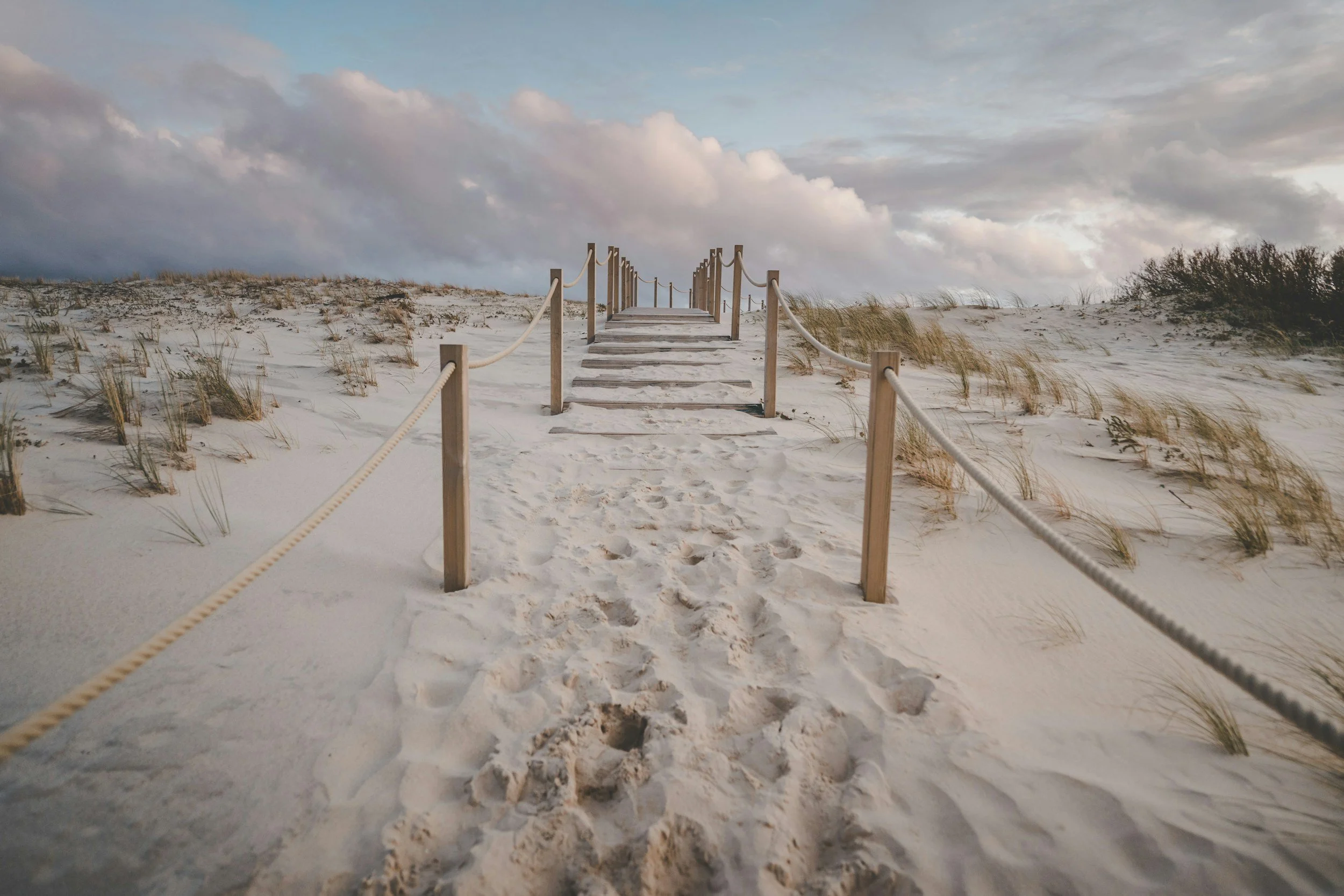 Wooden pathway with posts and ropes leading over sand dunes towards the beach with cloudy sky.