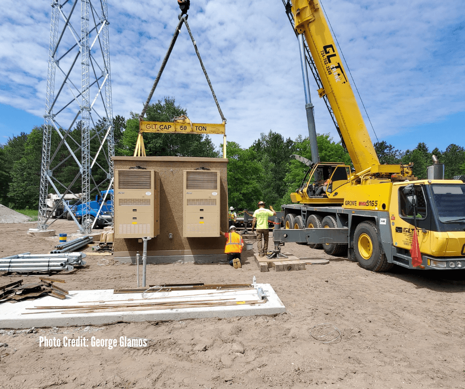 A yellow crane lifting a large electrical transformer at a construction site. Several workers are present, some observing and others working near the transformer. The background features construction equipment, a metal tower structure, trees, and a p