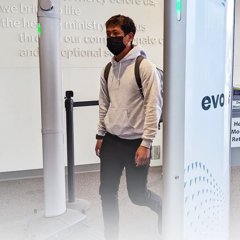 A young man wearing a black face mask, gray hoodie, and black pants, standing at an airport security checkpoint with a backpack on his shoulder. He is next to a white security scanner with the word 'evo' on it.
