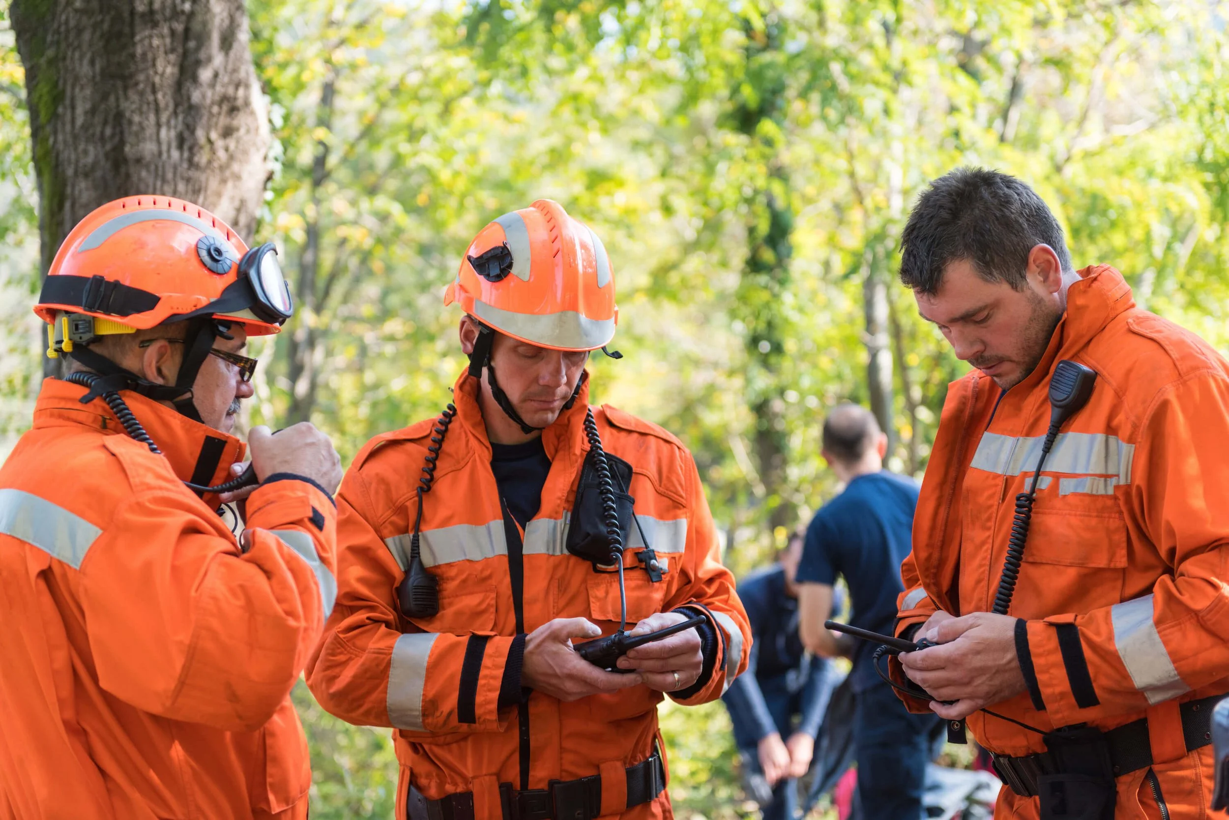 Three emergency rescue workers in orange uniforms and helmets with headlamps, standing outdoors in a wooded area, looking down at radios in their hands, with trees and a few other people in the background.