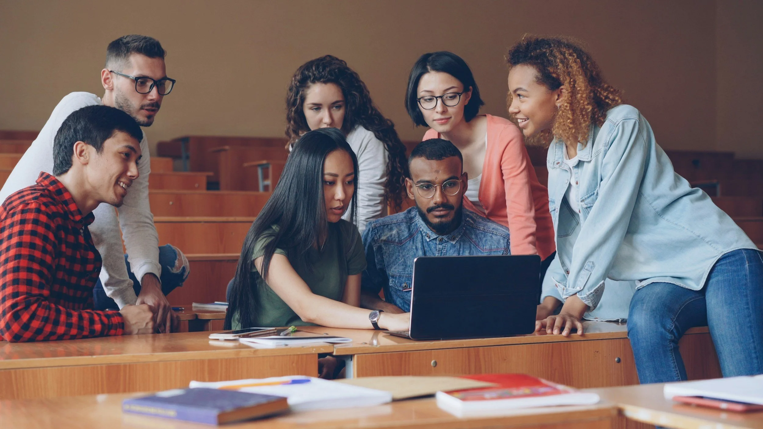 Group of seven students gathered around a laptop in a classroom, engaging in a discussion.
