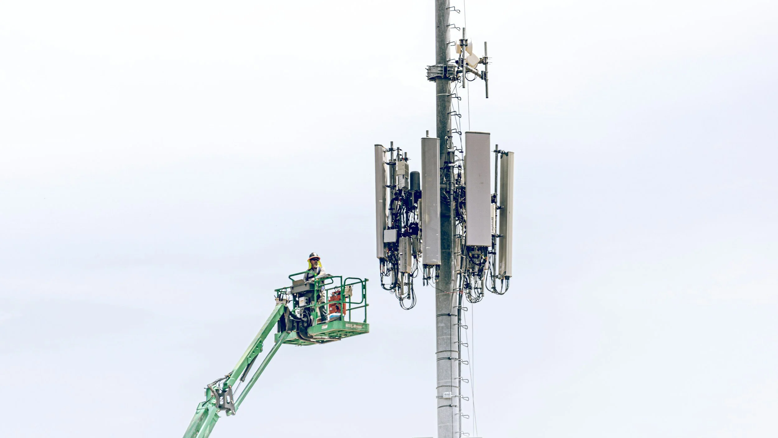 A worker on a green lift working on cellular antennas mounted on a tall utility pole against a cloudy sky.