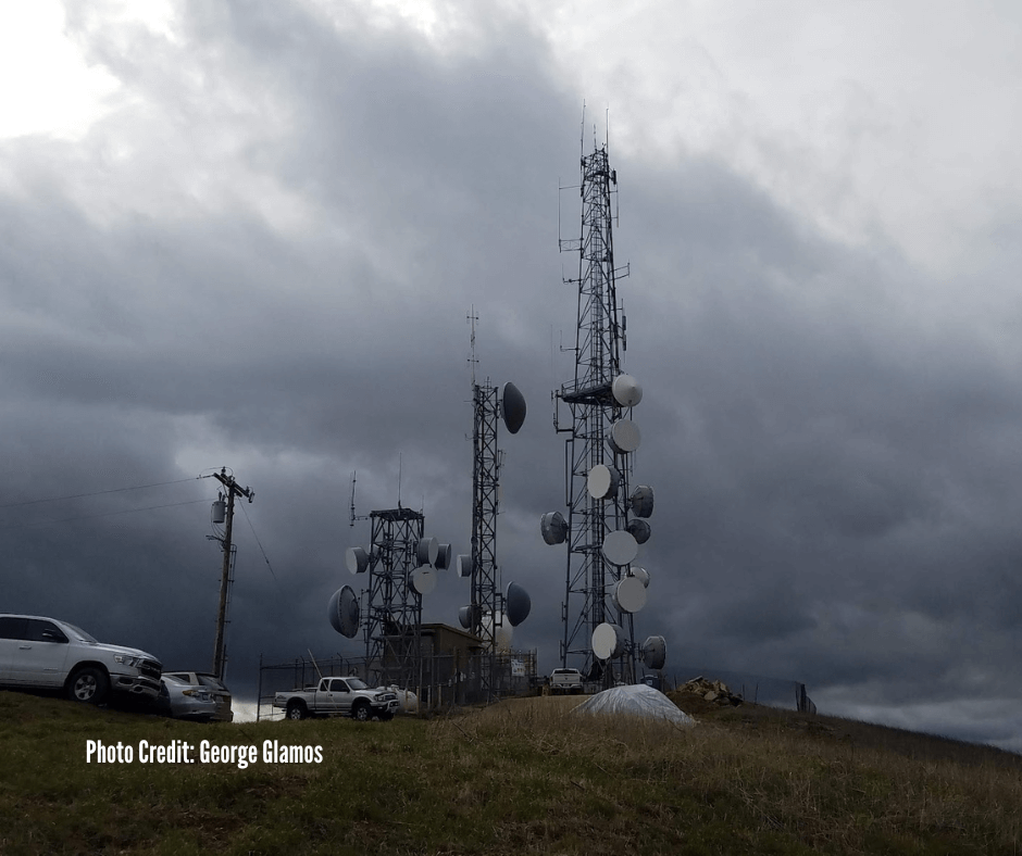 Cell towers with numerous antennas and dishes on a grassy hill under a cloudy sky, with parked cars nearby.