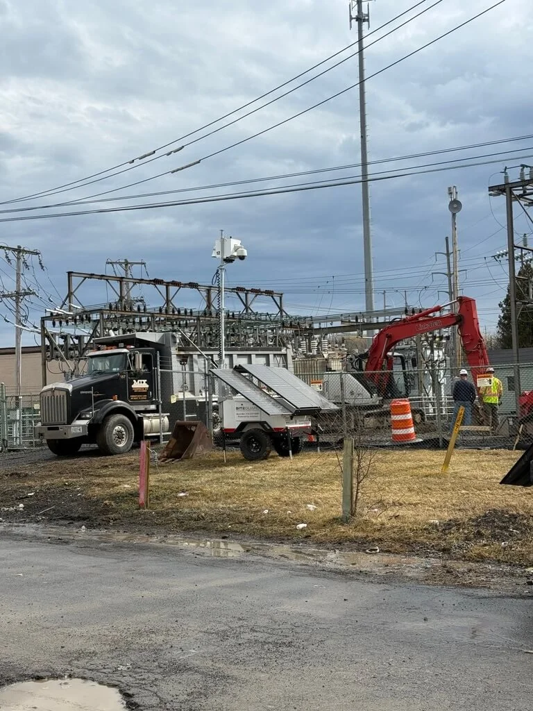 Workers with construction equipment, including a red excavator, near electrical substation with power lines, fence, and a cloudy sky.