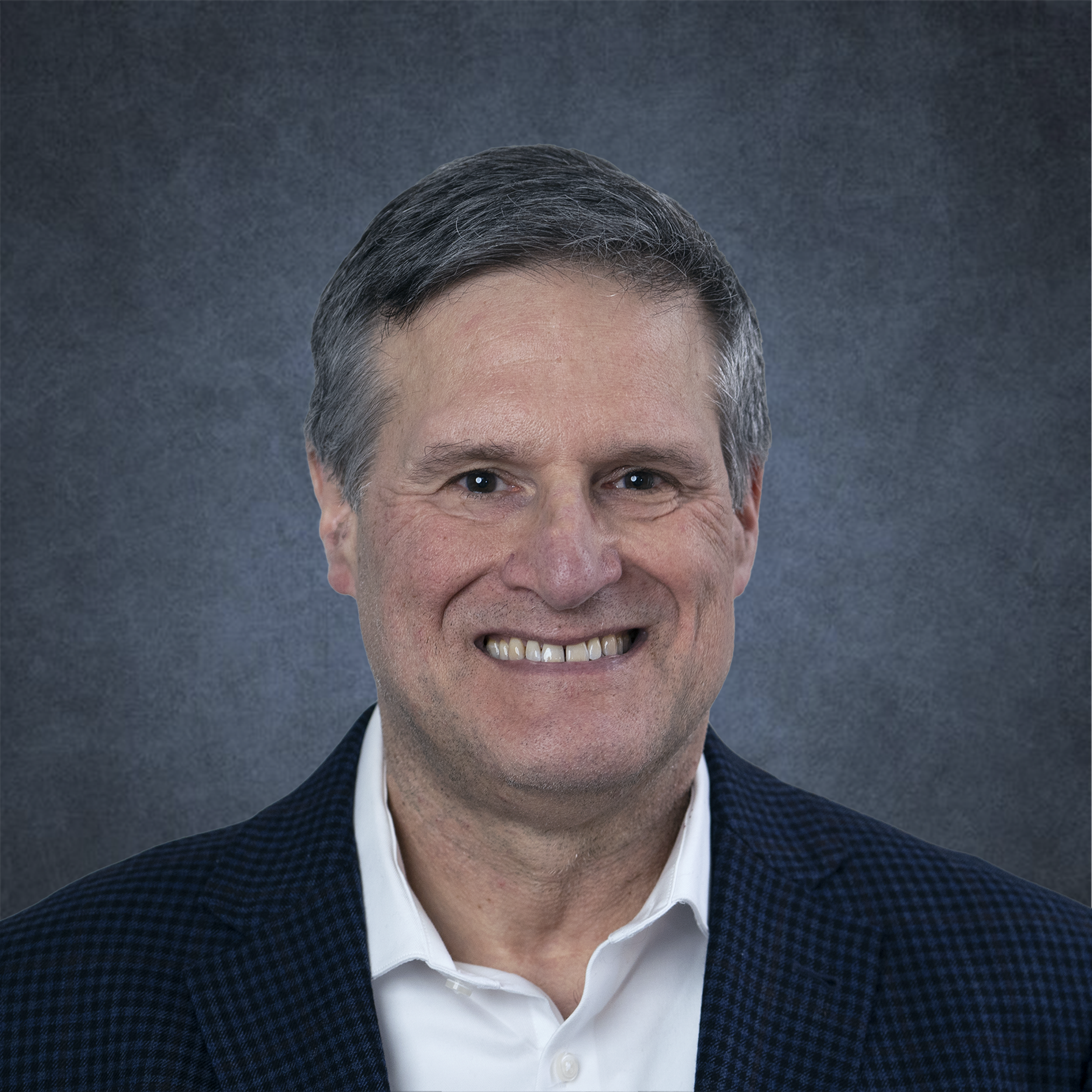 A middle-aged man with short, dark hair, wearing a dark blue checkered blazer and a white collared shirt, smiling at the camera against a textured gray background.