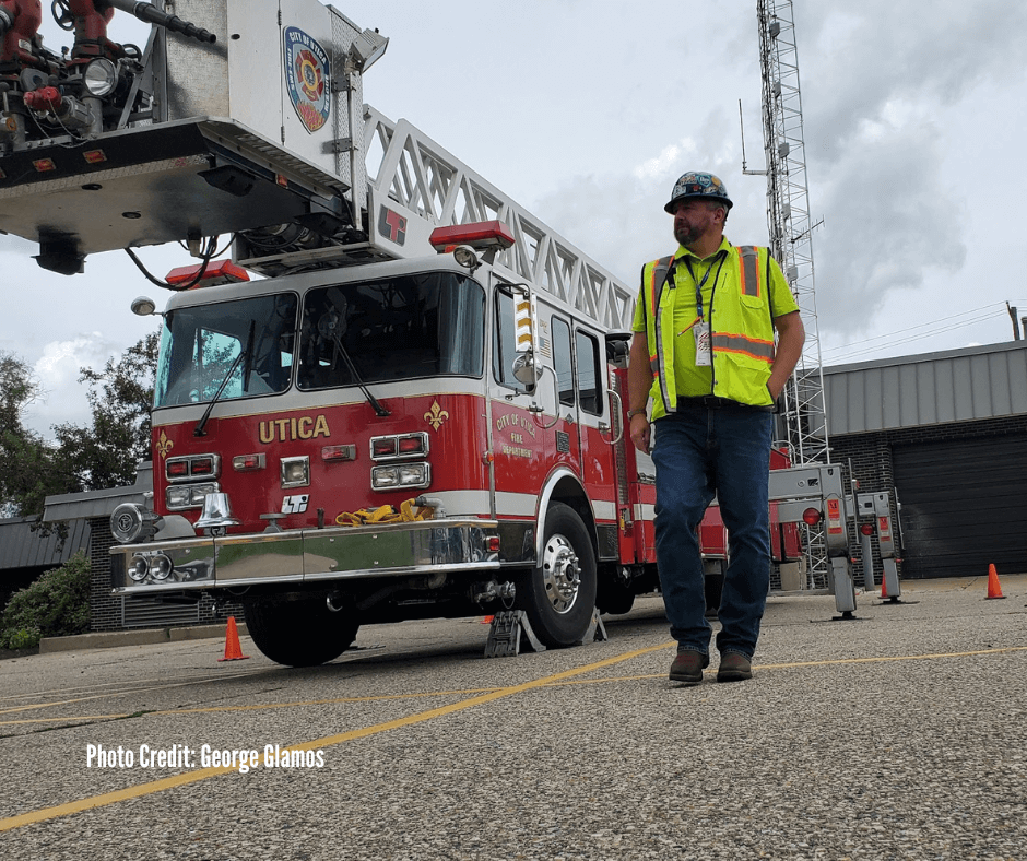 A firefighter standing next to a city of Utica fire truck with an extended ladder and a person wearing a yellow safety vest and helmet, during daytime with cloudy sky.