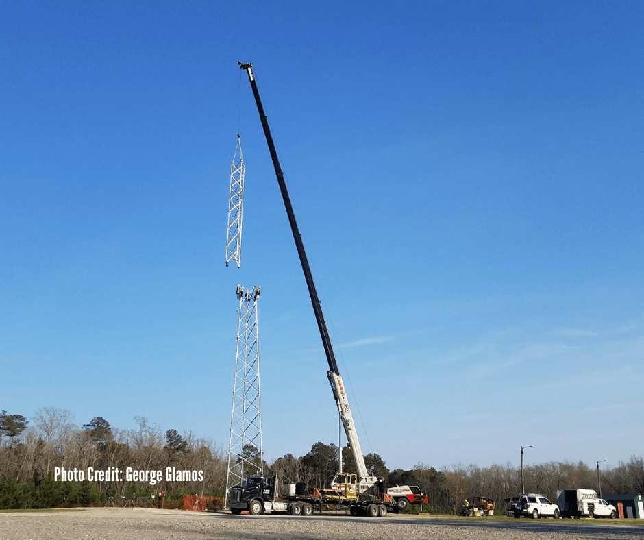 A crane lifting a large metal tower section into the air at an outdoor work site with several vehicles parked nearby and trees in the background.