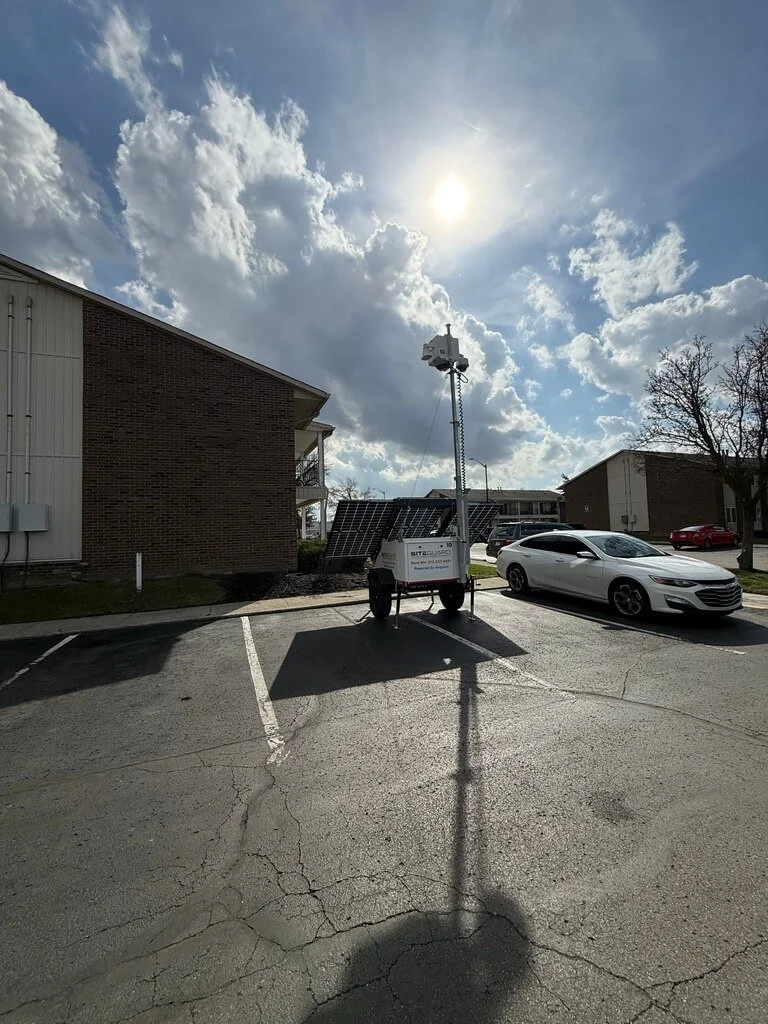 Parking lot with a solar-powered mobile weather station and solar panels, parked next to a white car, under a partly cloudy sky with the sun shining.