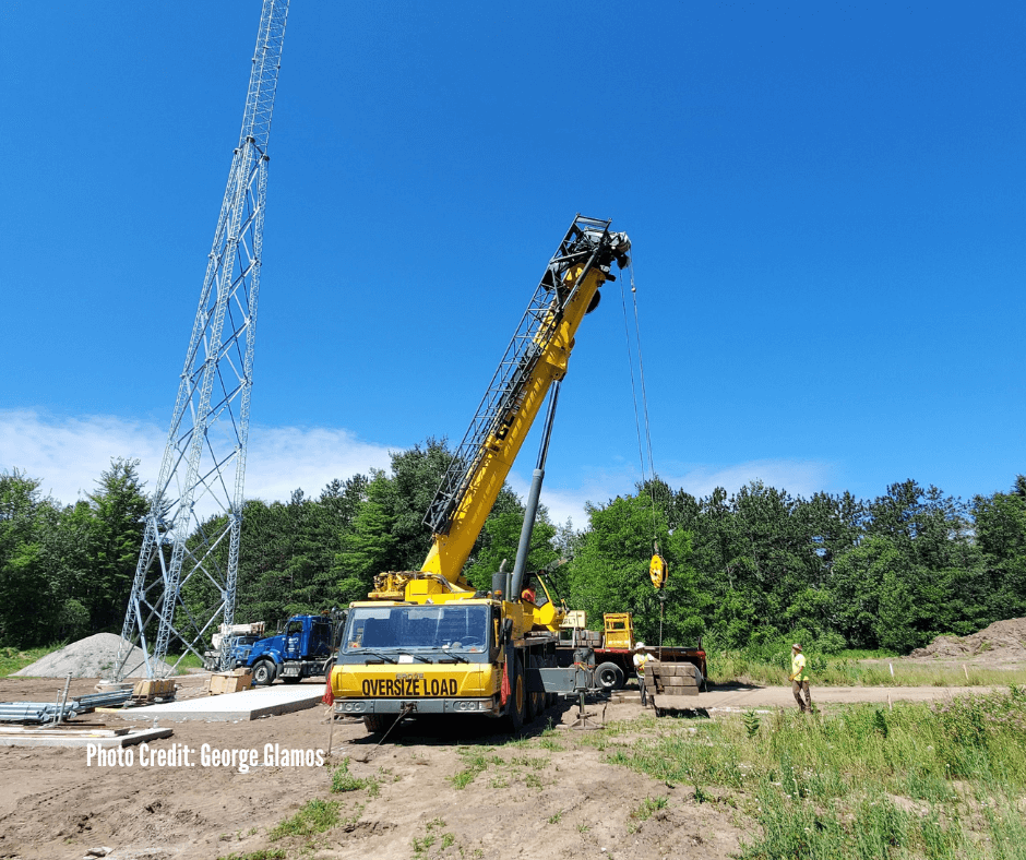 A construction site with a yellow crane lifting a large object, a tall communication tower, and workers nearby, set against a blue sky and green trees.