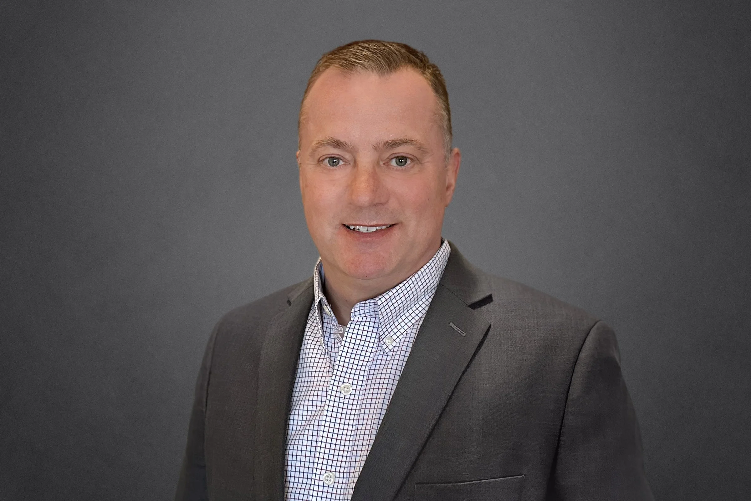 Professional headshot of a smiling man with short light brown hair, wearing a gray suit jacket and a white checkered shirt, against a dark gray background.