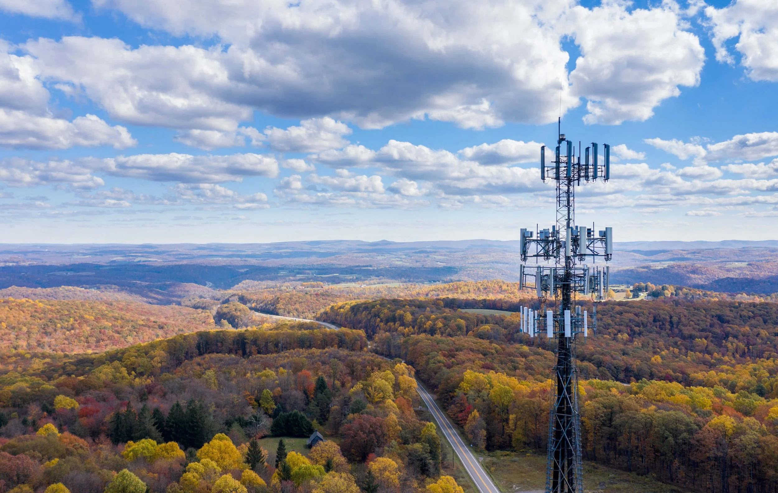 A tall telecommunications tower stands in a forested area with colorful autumn trees. A winding road runs through the landscape under a partly cloudy sky.