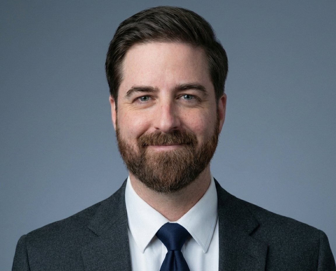 A portrait of a man with dark brown hair and a beard, wearing a dark suit, white shirt, and dark tie, smiling against a plain grey background.
