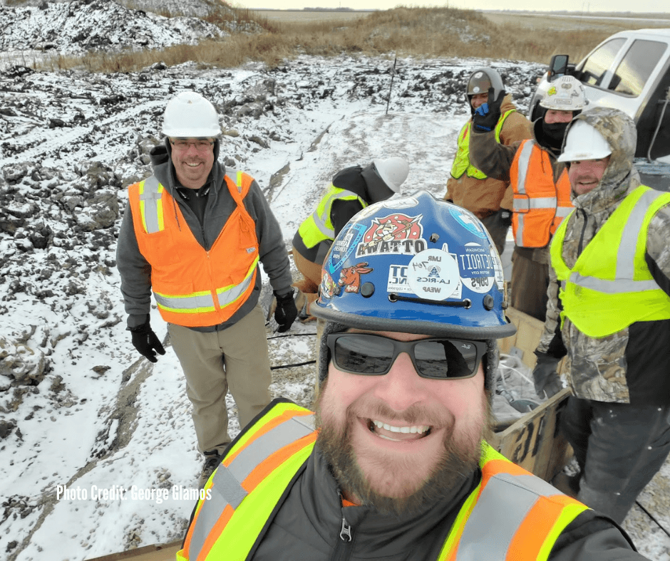 Group of construction workers smiling in snow, wearing safety vests and helmets, with some working on a project outdoors.