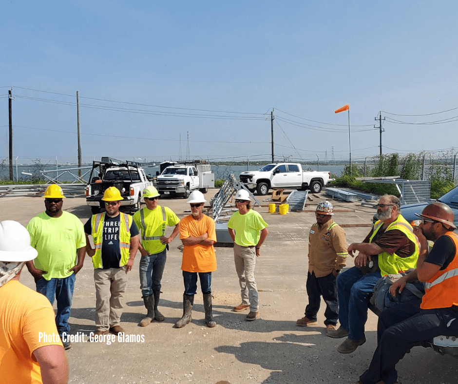 Group of construction workers standing together outdoors at a construction site, wearing safety helmets and reflective vests, with trucks and construction materials in the background.
