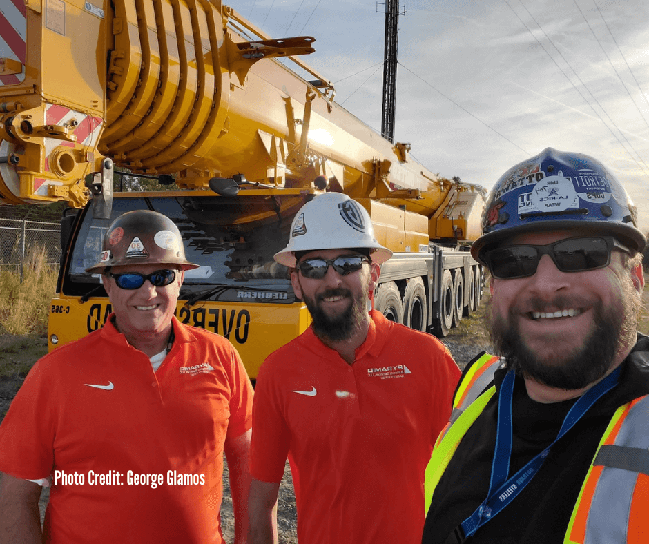 Three men in construction helmets and safety vests posing in front of a large yellow crane truck.