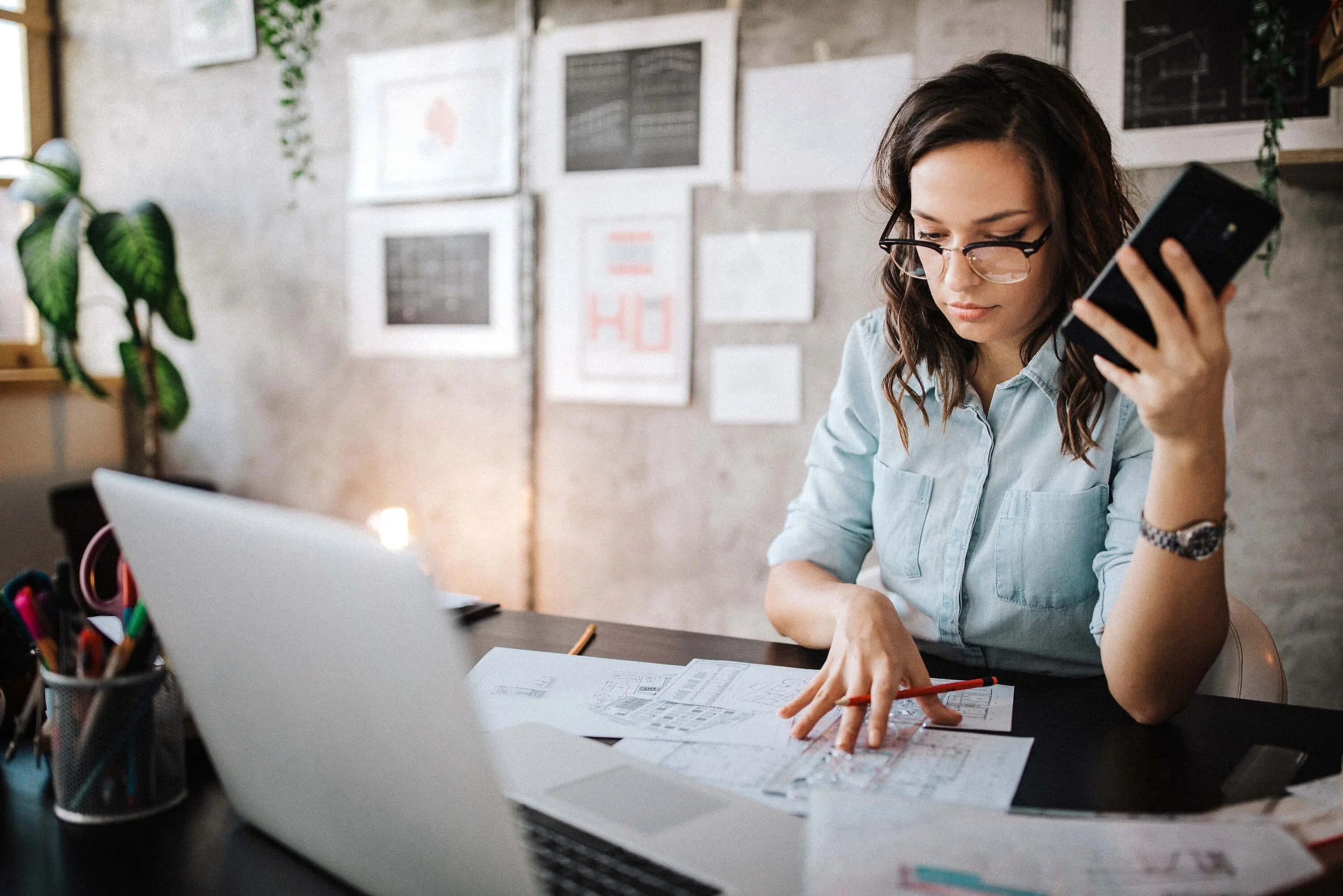 A woman works at her desk with a laptop computer, cell phone, and project drawing blueprints.