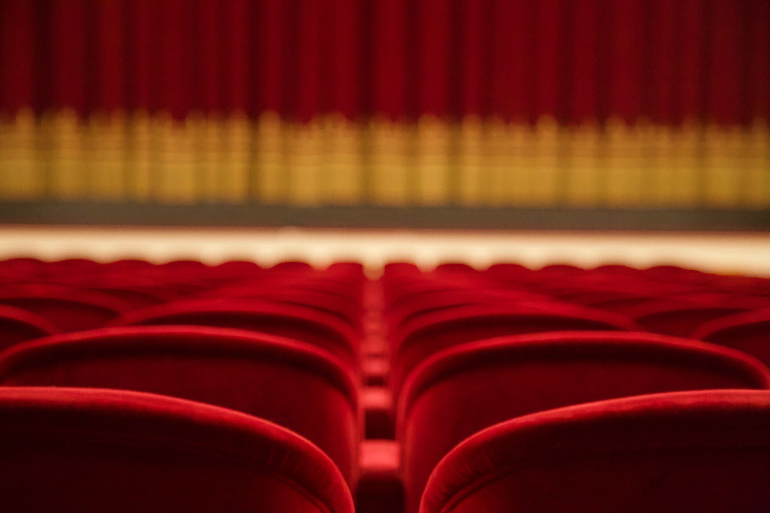 View from the back of red velvet theater seats facing a gold and red curtain.