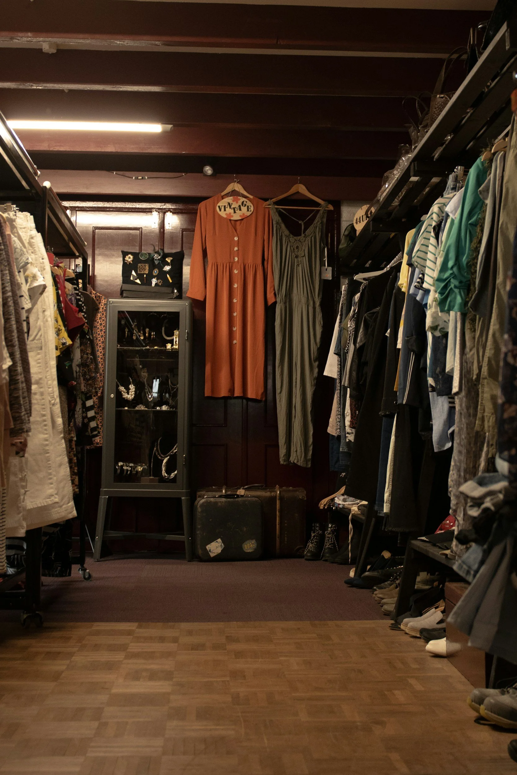 A vintage clothing and accessories store with racks of clothes on both sides, a display case with jewelry in the center, and wooden walls. A long orange dress and a green dress are hanging on the back wall. The store has wood flooring and warm lighting.