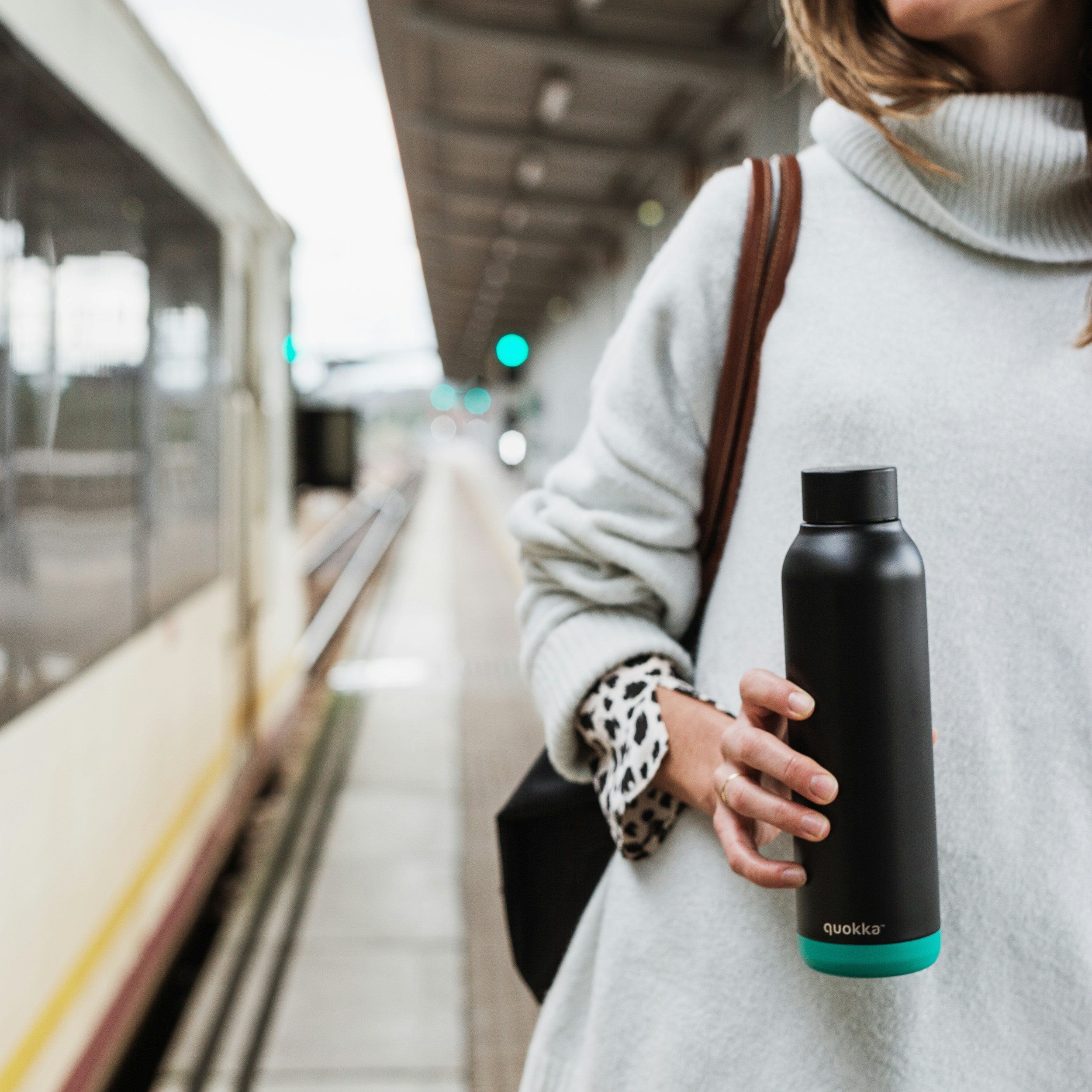 Close-up of a person wearing a white sweater with leopard print cuffs, holding a black insulated water bottle with a teal bottom, at a train station platform with a train and green signal in the background.