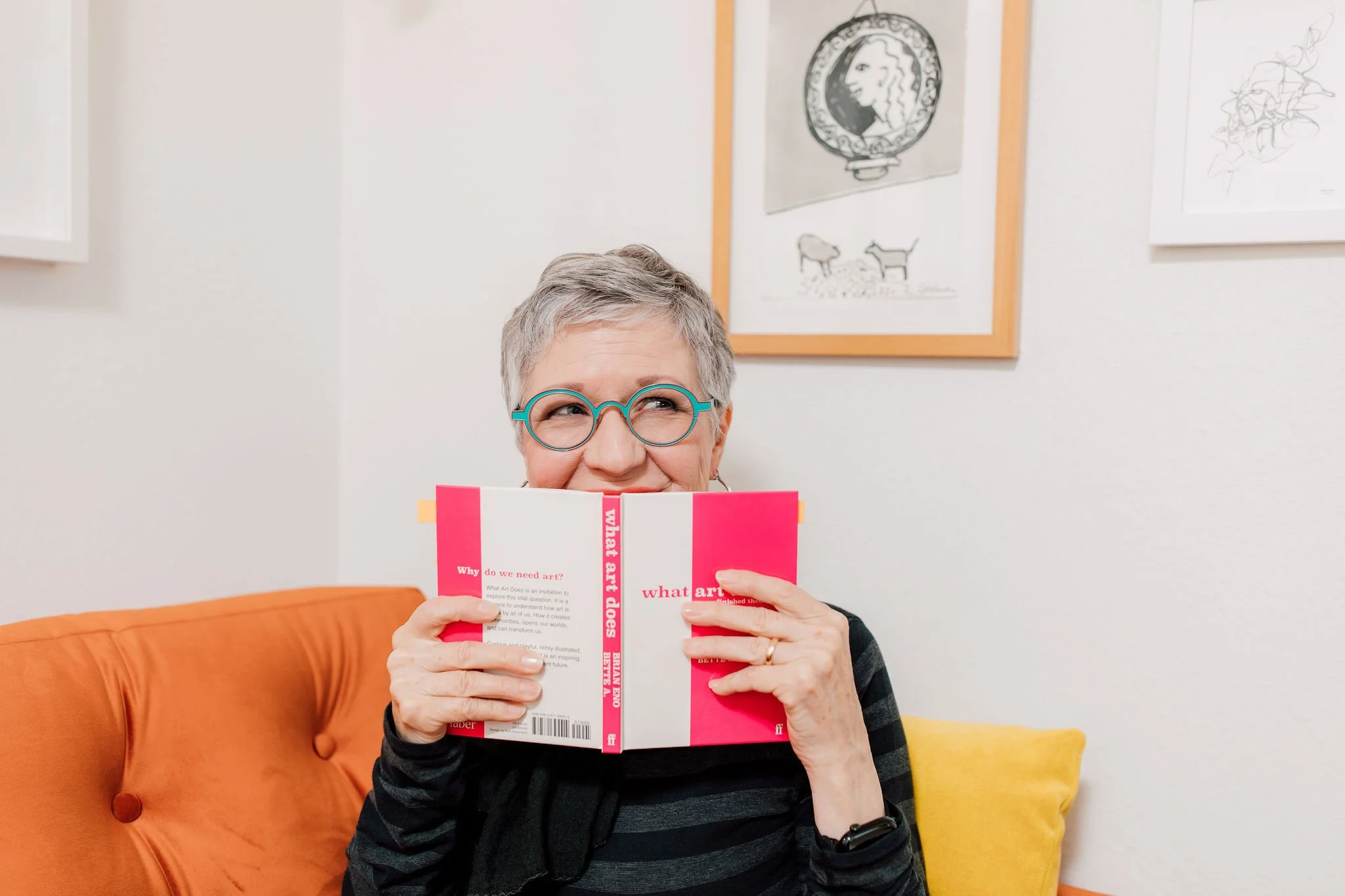 A woman with gray hair and glasses holding a red and white book titled 'What Art Does' in front of her face, with a colorful couch and framed artwork in the background.