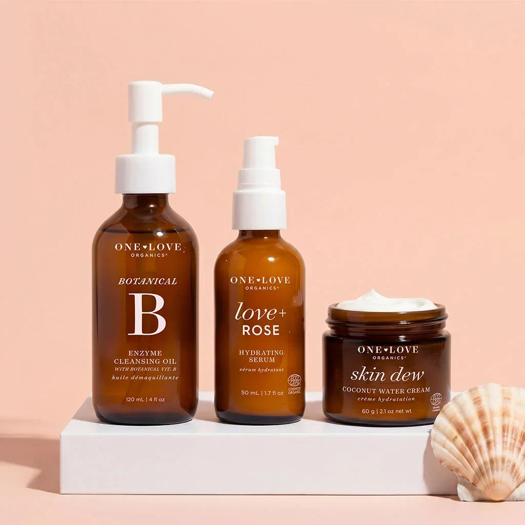 Three amber skincare products from One Love Organics on a white platform with a shell, set against a pink background. The products include enzyme cleansing oil, hydrating serum, and coconut water cream.