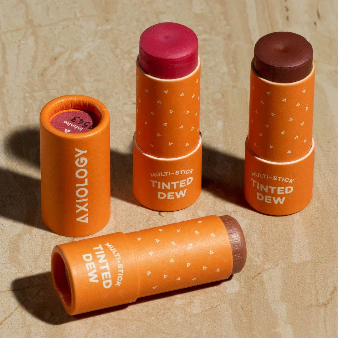 Four orange multi-stick tinted lip balms on a beige surface, three standing upright and one lying down, with visible colored tops in shades of red and brown, and white text on the packaging.
