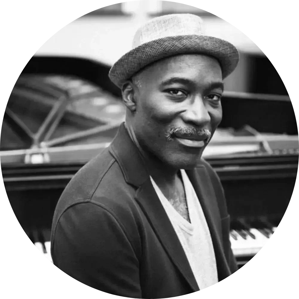 A black and white photo of a man with a mustache and wearing a fedora hat, standing in front of a piano, smiling at the camera.