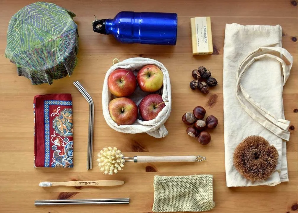 Eco-friendly shopping essentials including a reusable net bag, stainless steel straw, soap, reusable tote, apples, dried fruits, a metal skewer, reusable cleaning brush, bandana, eco-friendly wool brush, and cloths on a wooden surface.